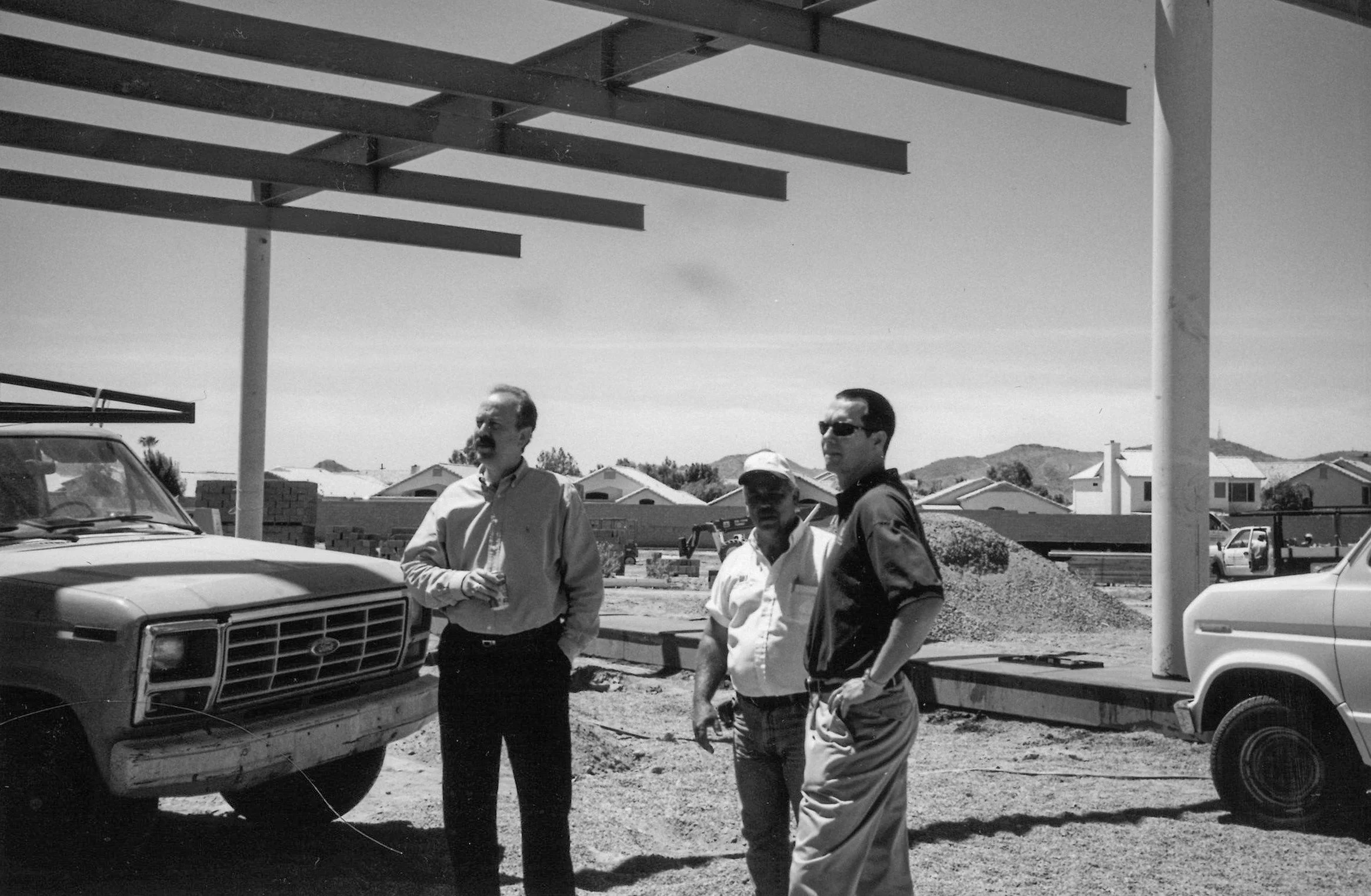 Three men standing outdoors at a construction site near parked trucks, with houses and hills in the background.