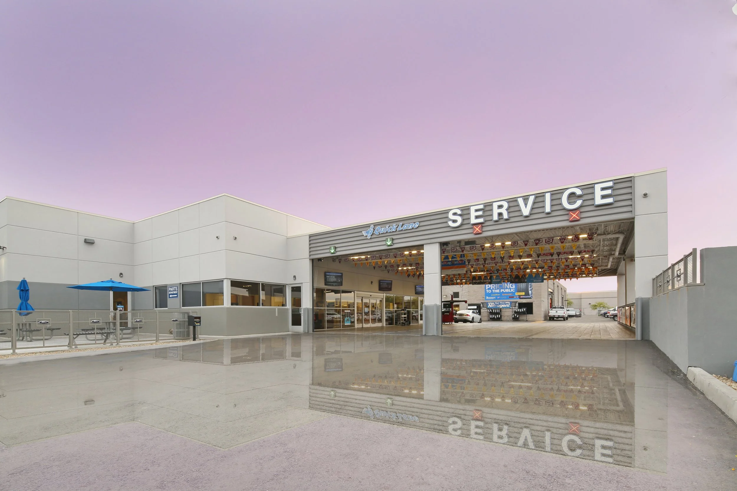 Auto service center with a large sign reading 'SERVICE', a small advertising board, cars parked inside, a patio with tables and umbrella on the left, and a purple sky in the background.