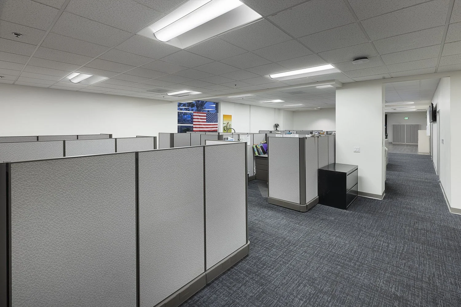 Empty office cubicles with gray partitions, a window with an American flag, and a hallway leading to a door.