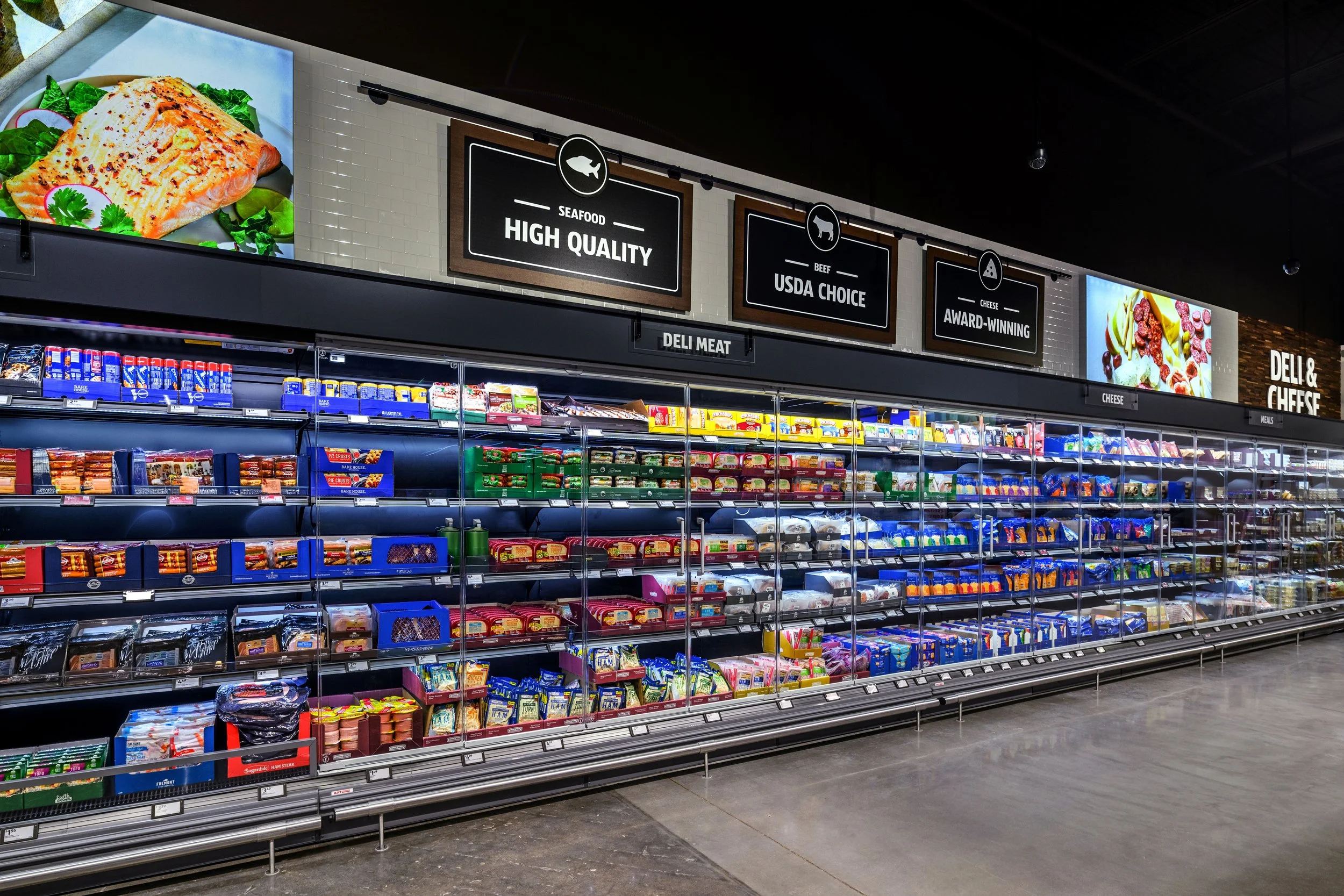 Grocery store refrigerated section with shelves of packaged foods, signage for seafood, beef, cheese, and deli meat.