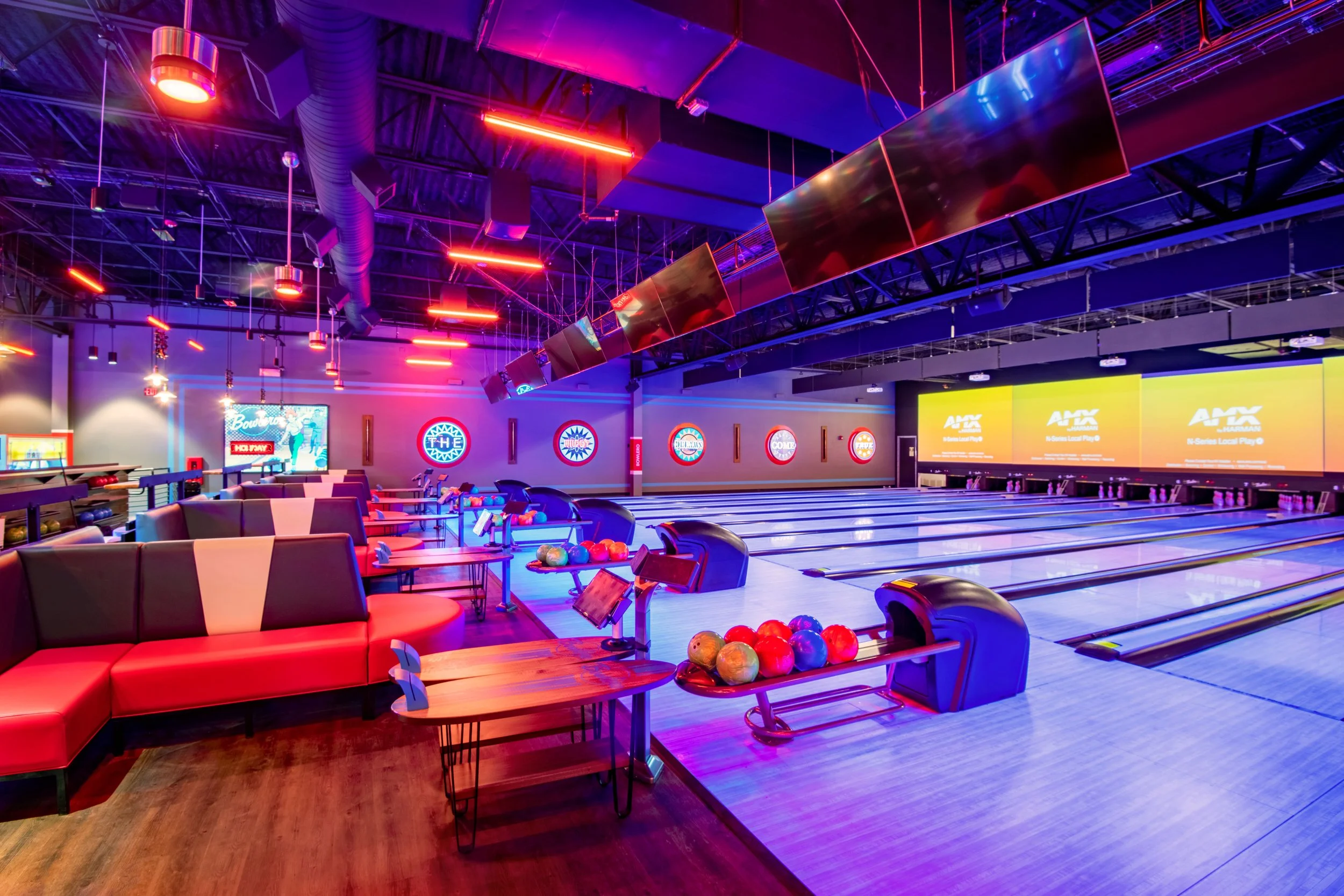 Interior of a modern bowling alley with multiple lanes, colorful neon lighting, digital screens, and seating area with red and black couches.