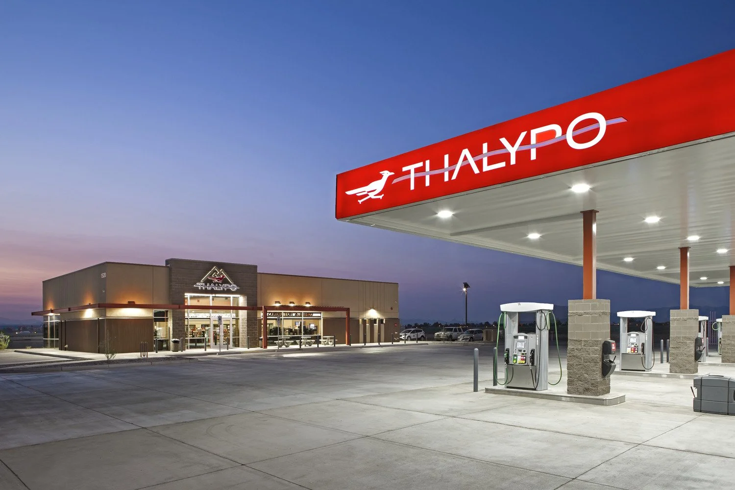 A Thaiyum convenience store and gas station at dusk with a four-pump fuel island and a building with a dark brick facade and illuminated sign.