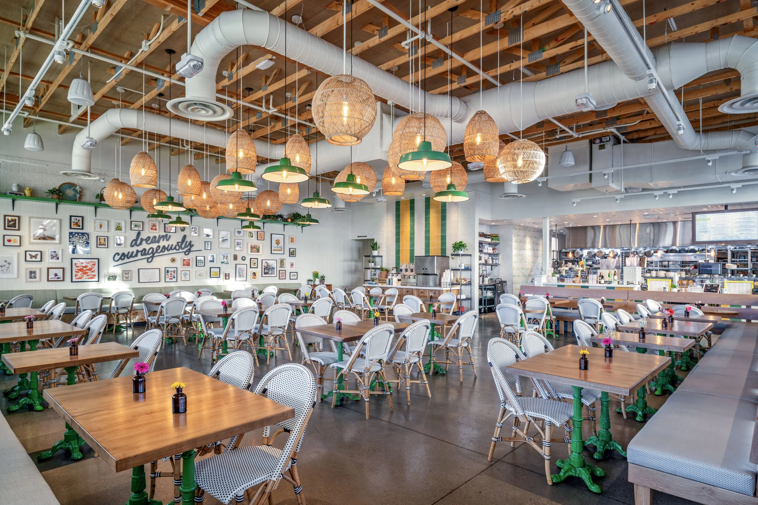 Brightly lit cafe with wooden tables, white chairs with black and white striped cushions, and hanging wicker and green-shaded lamps. Decor includes framed pictures and a wall with the slogan 'dream courageously'.