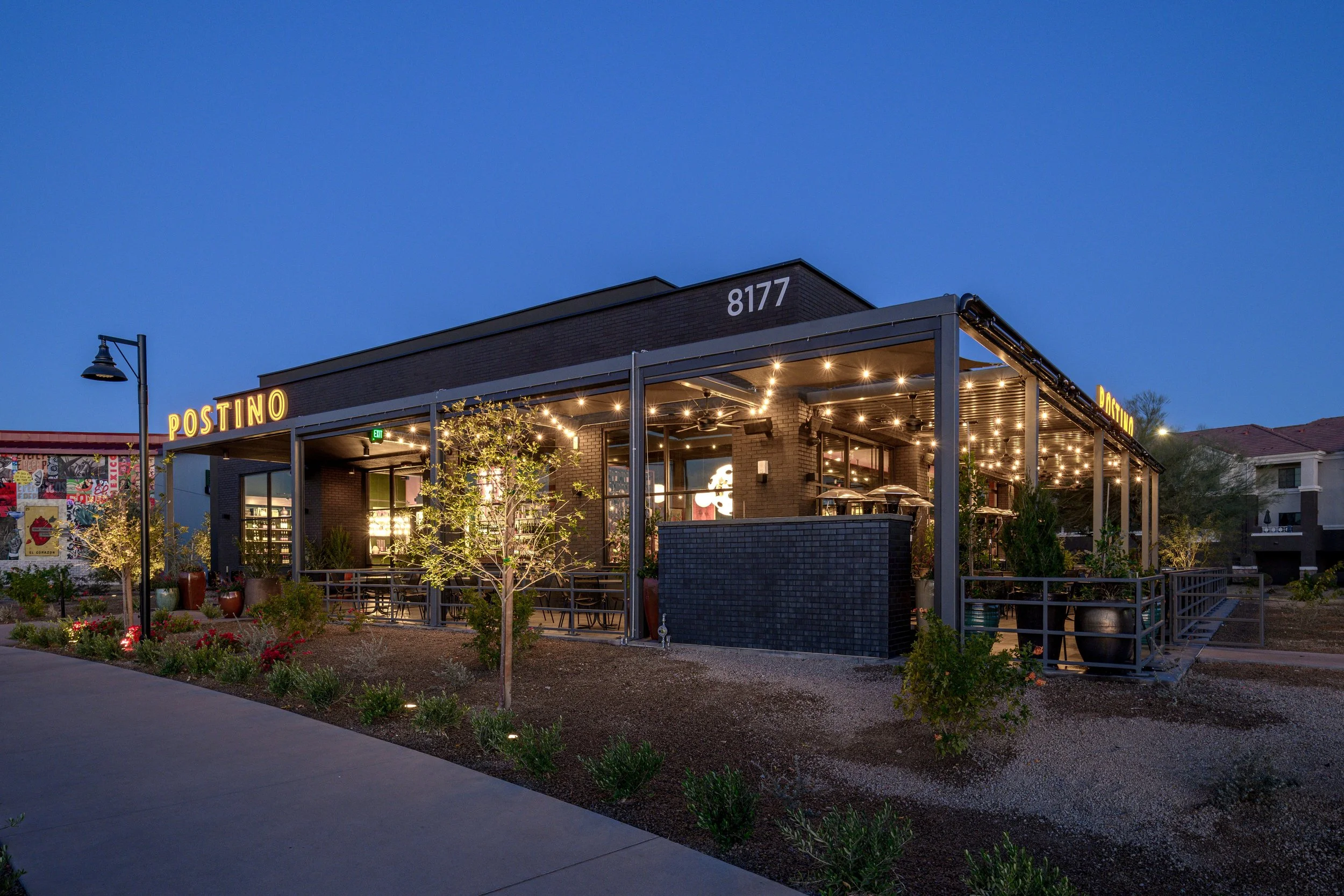 Exterior view of Postino restaurant during evening with warm lighting, outdoor seating, and landscaping.