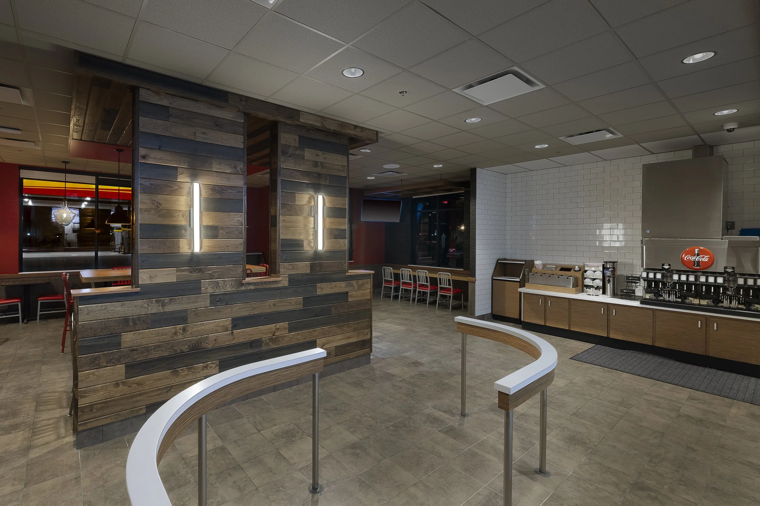 Empty fast food restaurant dining area with a self-serve drink station, including a Coca-Cola soda dispenser, surrounded by wooden and metal furniture, with tiled floors and ceiling, and a view of some chairs and tables.