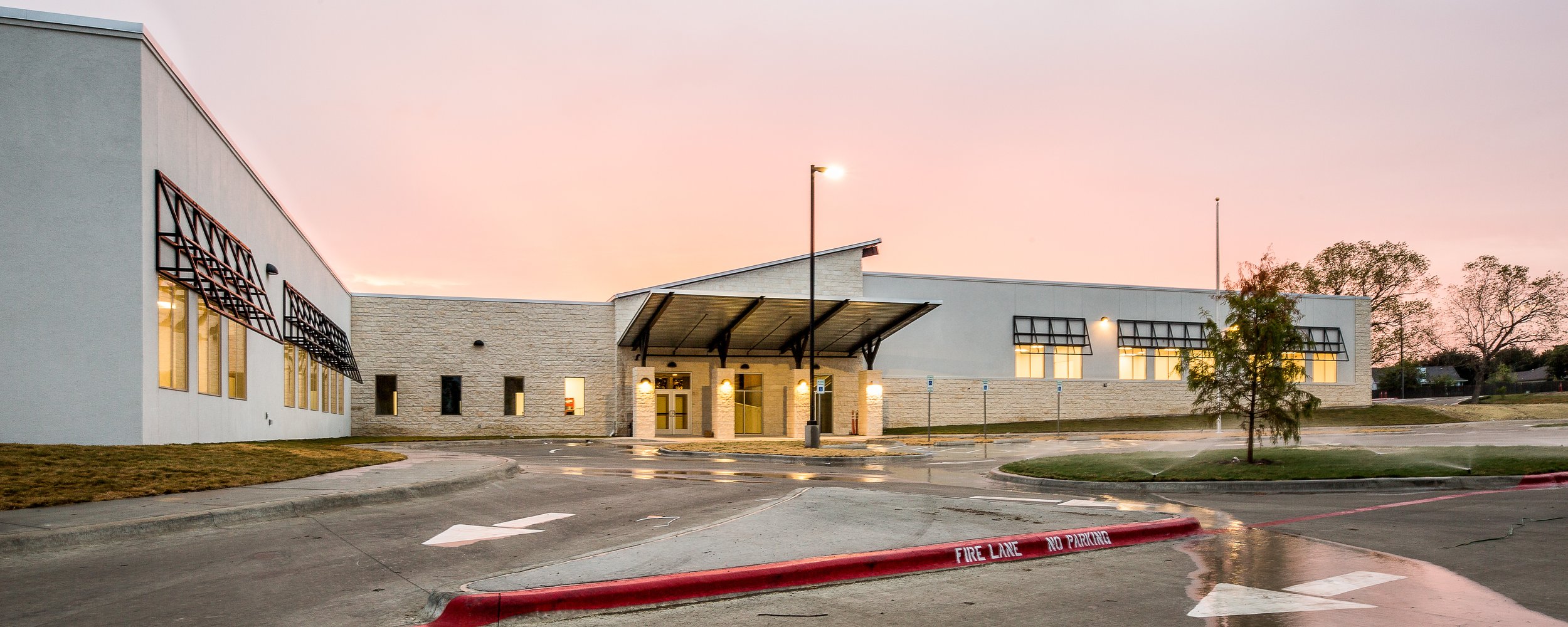 Modern commercial building with a parking lot, illuminated at dusk, with a pink sky and trees in the background.