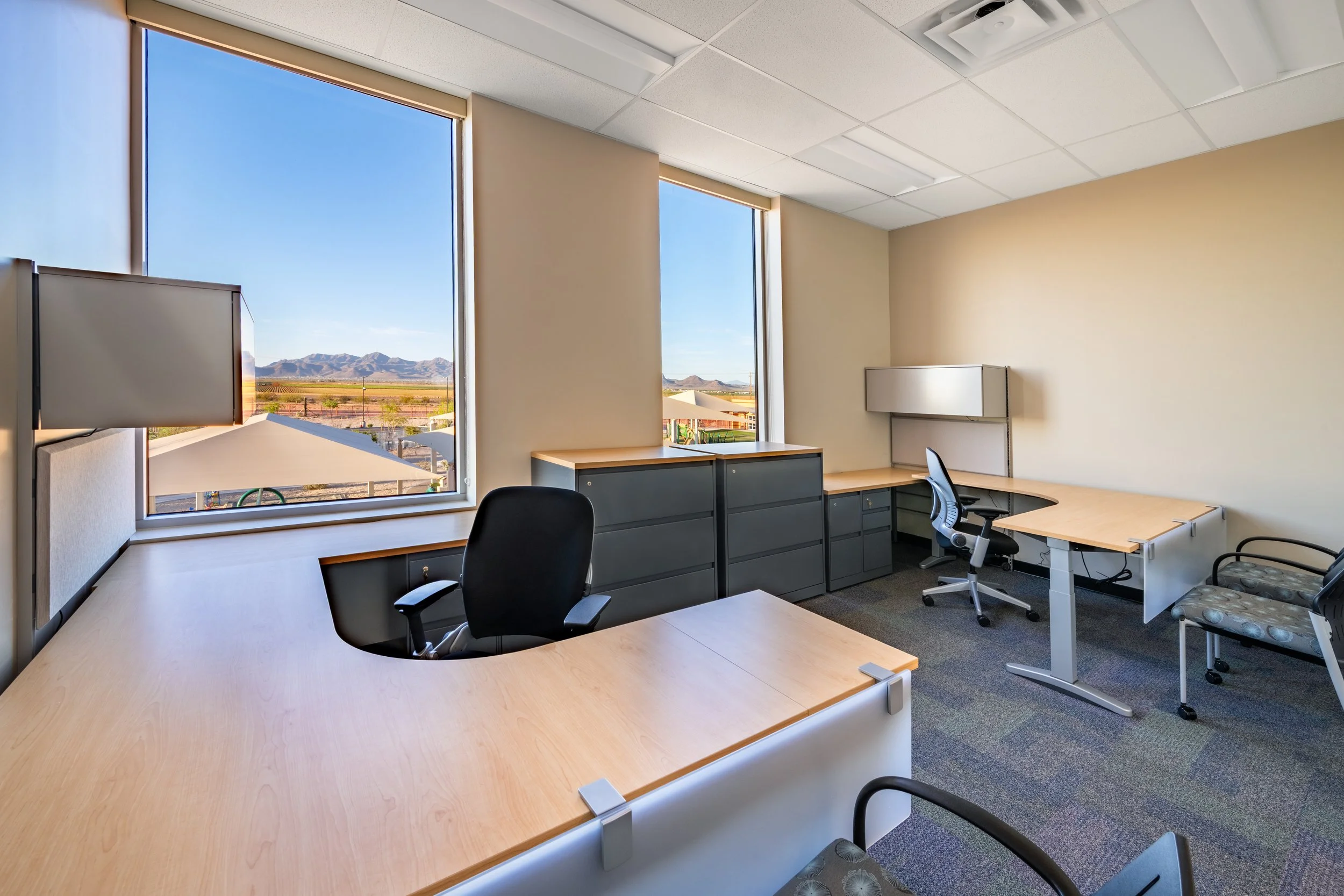 Office space with two large windows showing mountains and desert landscape, L-shaped wooden desk, black office chair, gray filing cabinets, small side table, gray wall-mounted shelf, hanging monitor, beige walls, ceiling tiles, and gray carpet.