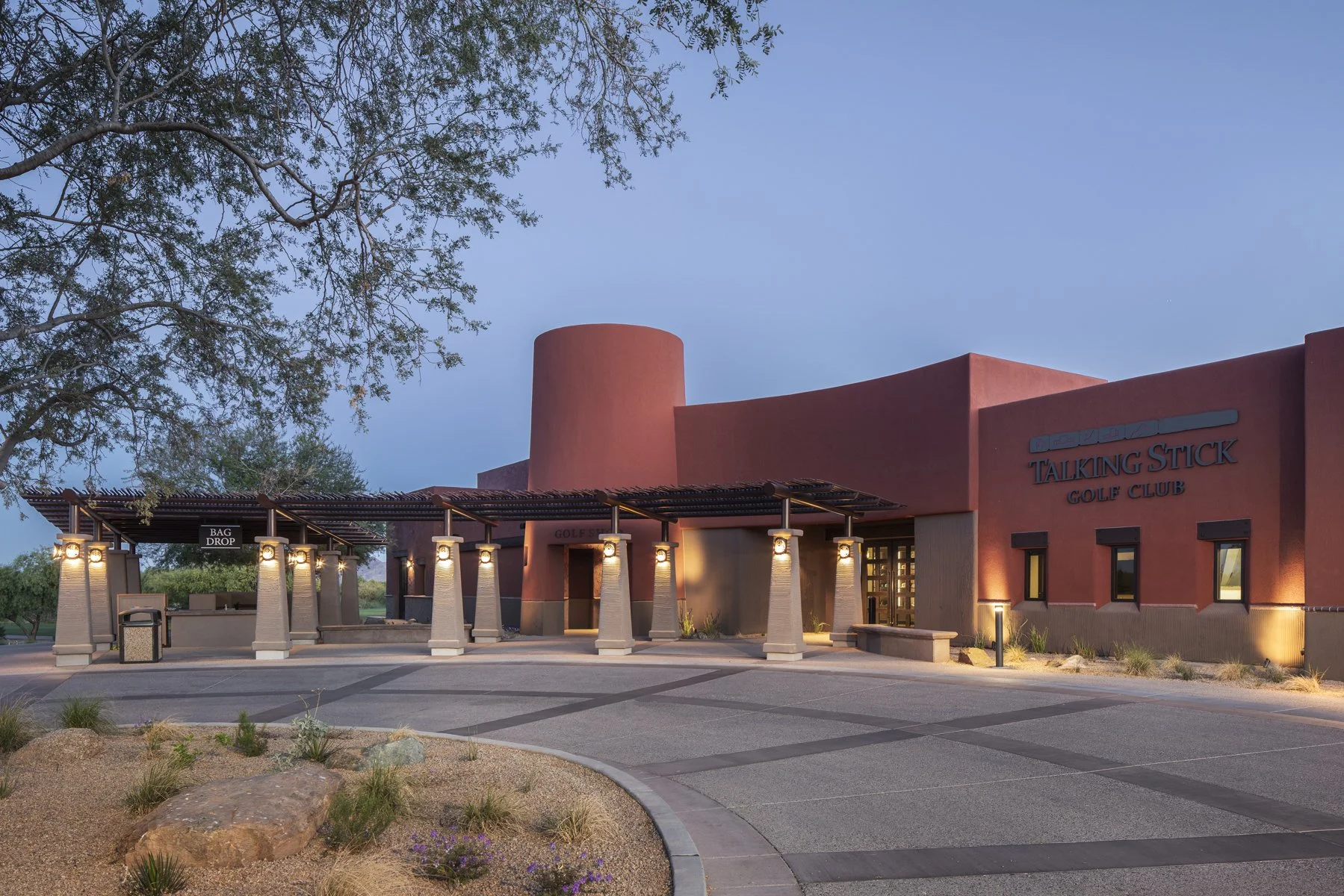 Exterior view of the Talking Stick Golf Club at dusk, with a modern, Adobe-style building, illuminated pathway, and landscaped desert area in the foreground.