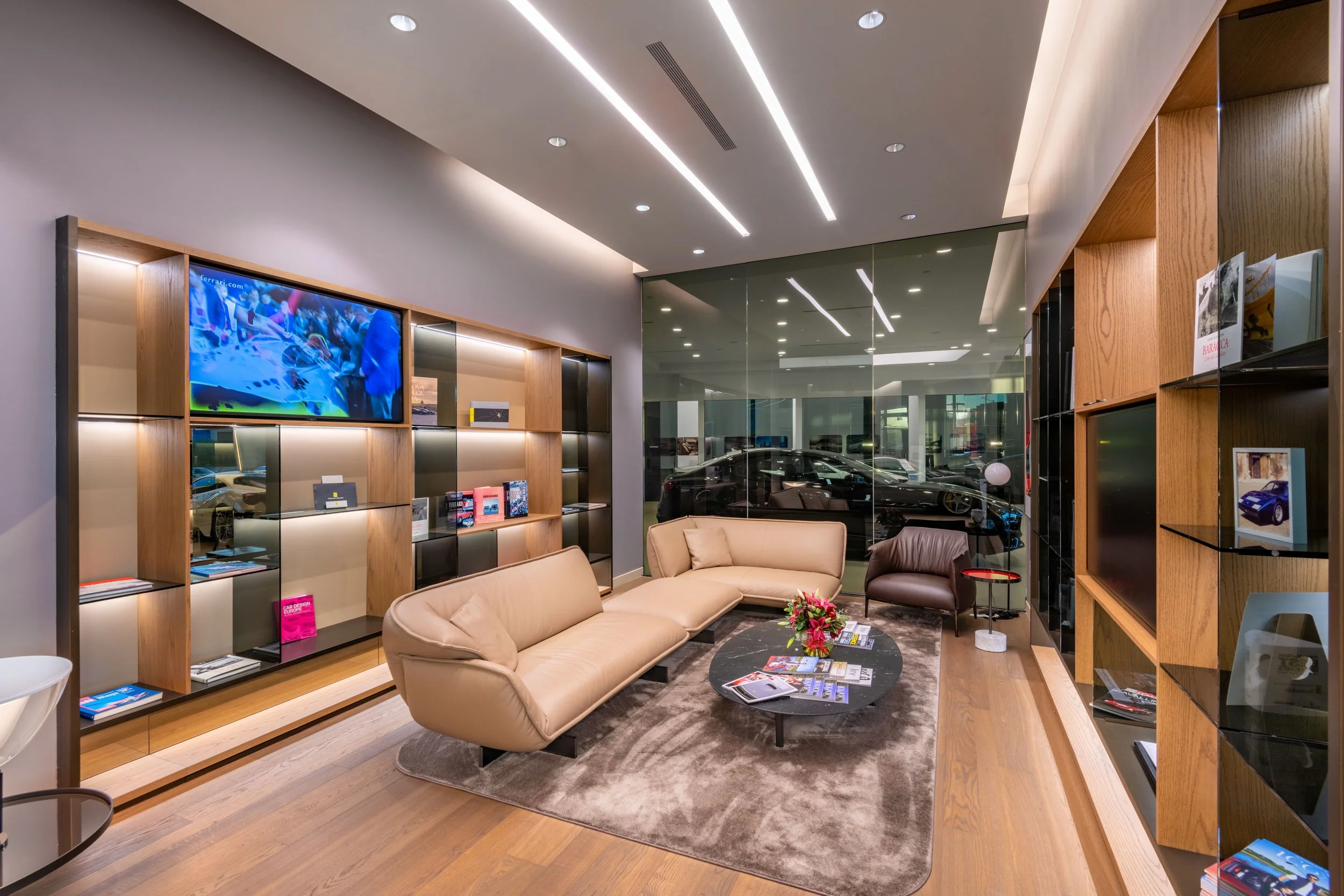 Luxury car showroom waiting area with beige sofas, a black coffee table, magazine brochures, a vase of flowers, a glass wall showing parked sports cars outside, and wooden bookshelves with magazines, brochures, and a television.
