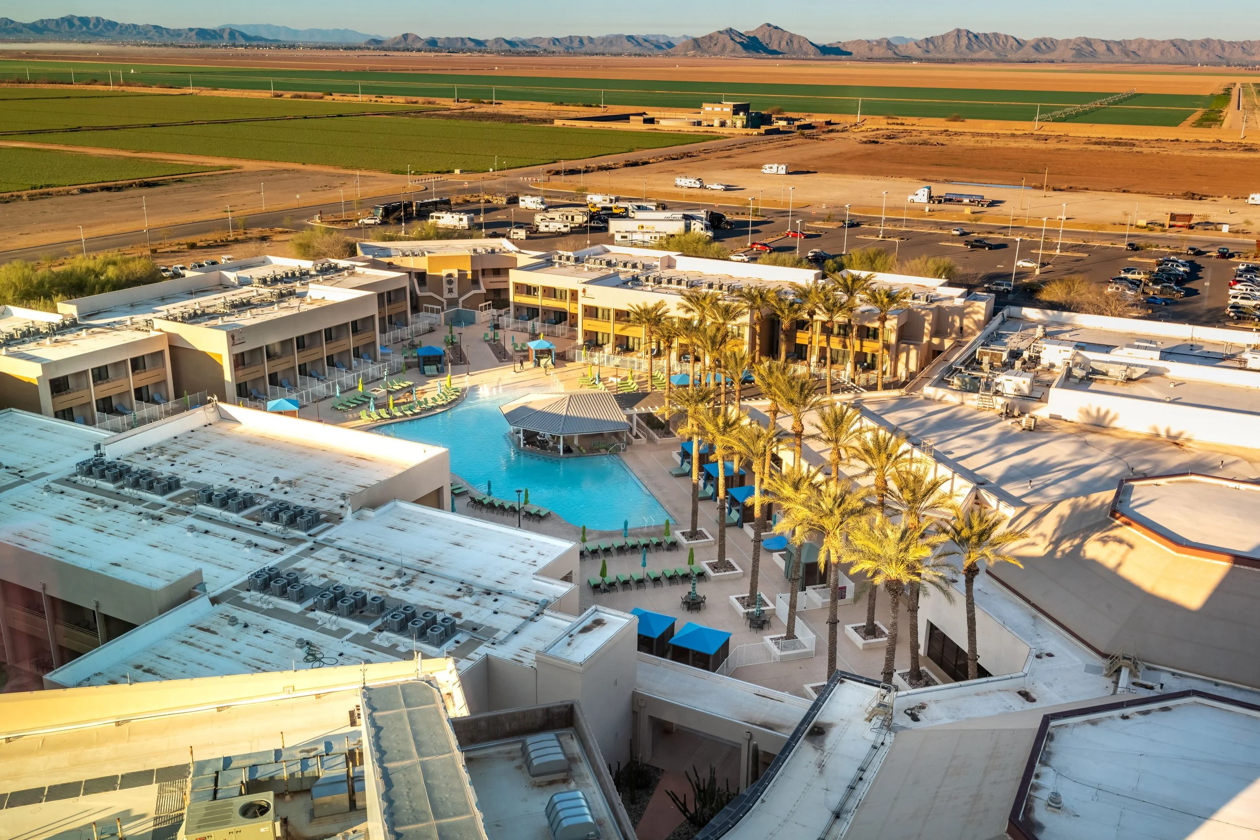 Aerial view of a hotel complex with a swimming pool surrounded by palm trees, lounge chairs, and shaded umbrellas, with parking lot and open desert landscape with mountains in the background.