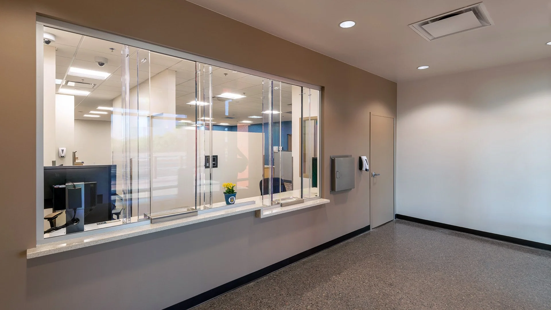 Empty hospital reception area with a windowed partition, a computer on the counter, a small potted plant, and a door on the right wall.
