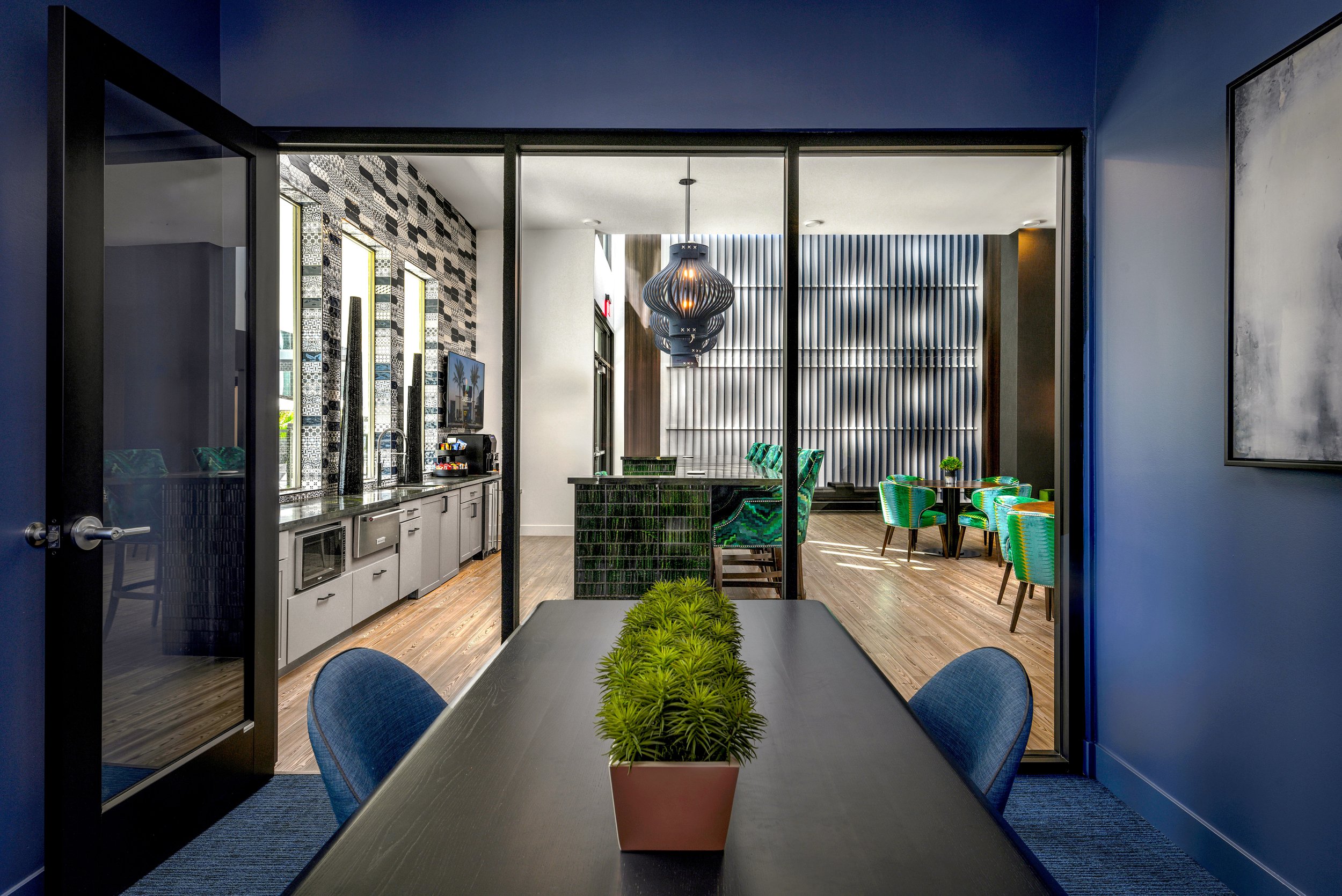 A modern indoor lounge area with a dining table, green chairs, and a decorative wall, viewed through glass doors from a room with a black table and blue chairs.