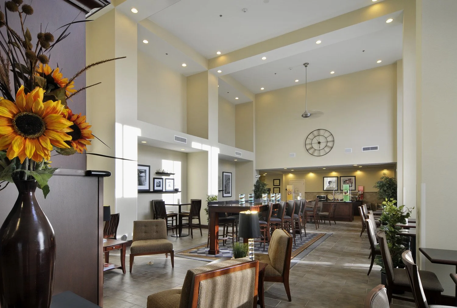 Interior of a modern lounge or lobby with high ceilings, large wall clock, and various seating areas including armchairs, dining chairs, and tables, decorated with plants and framed pictures.