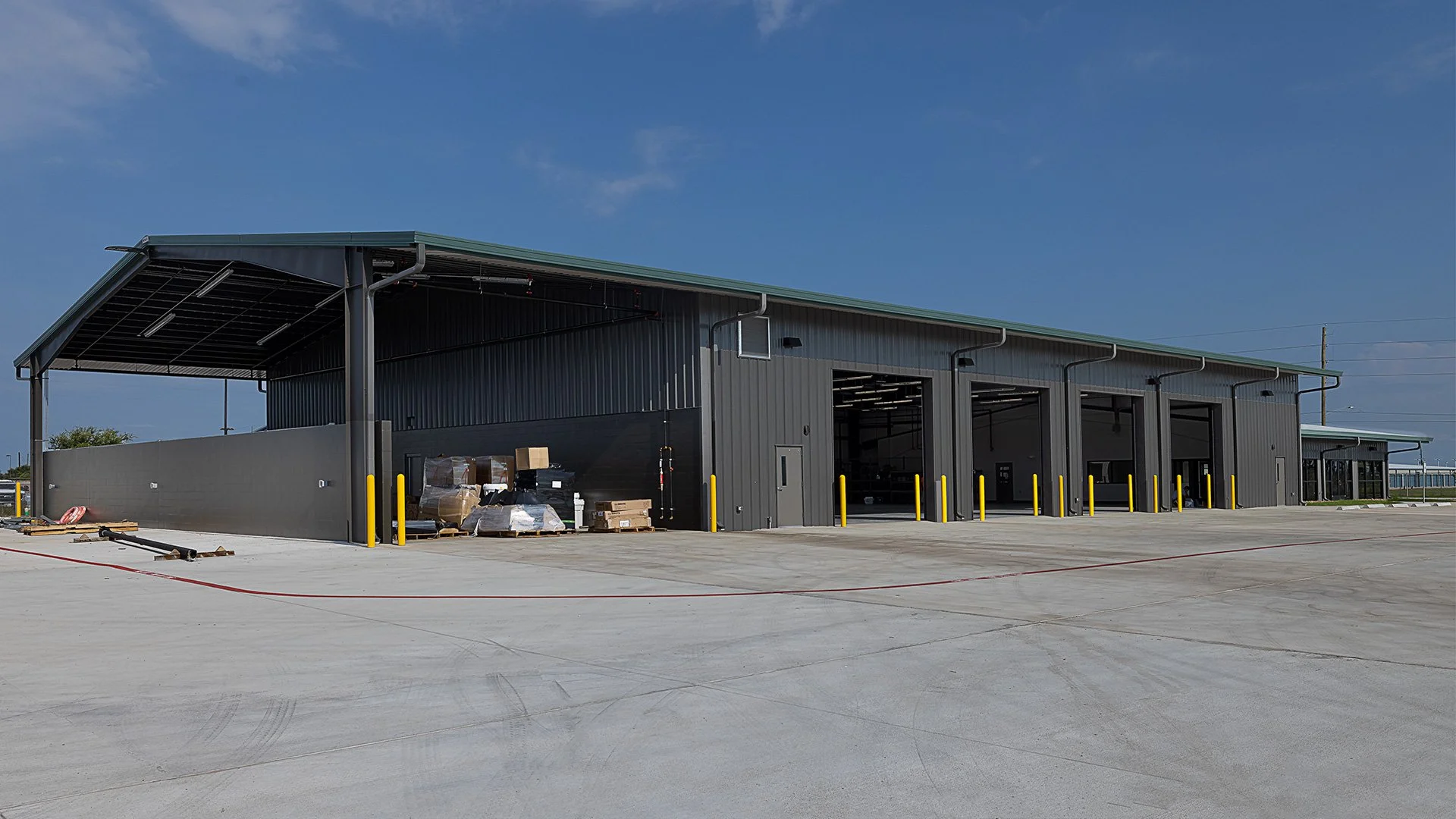 Newly constructed industrial warehouse building with large garage doors and loading area, surrounded by a concrete yard and clear blue sky.