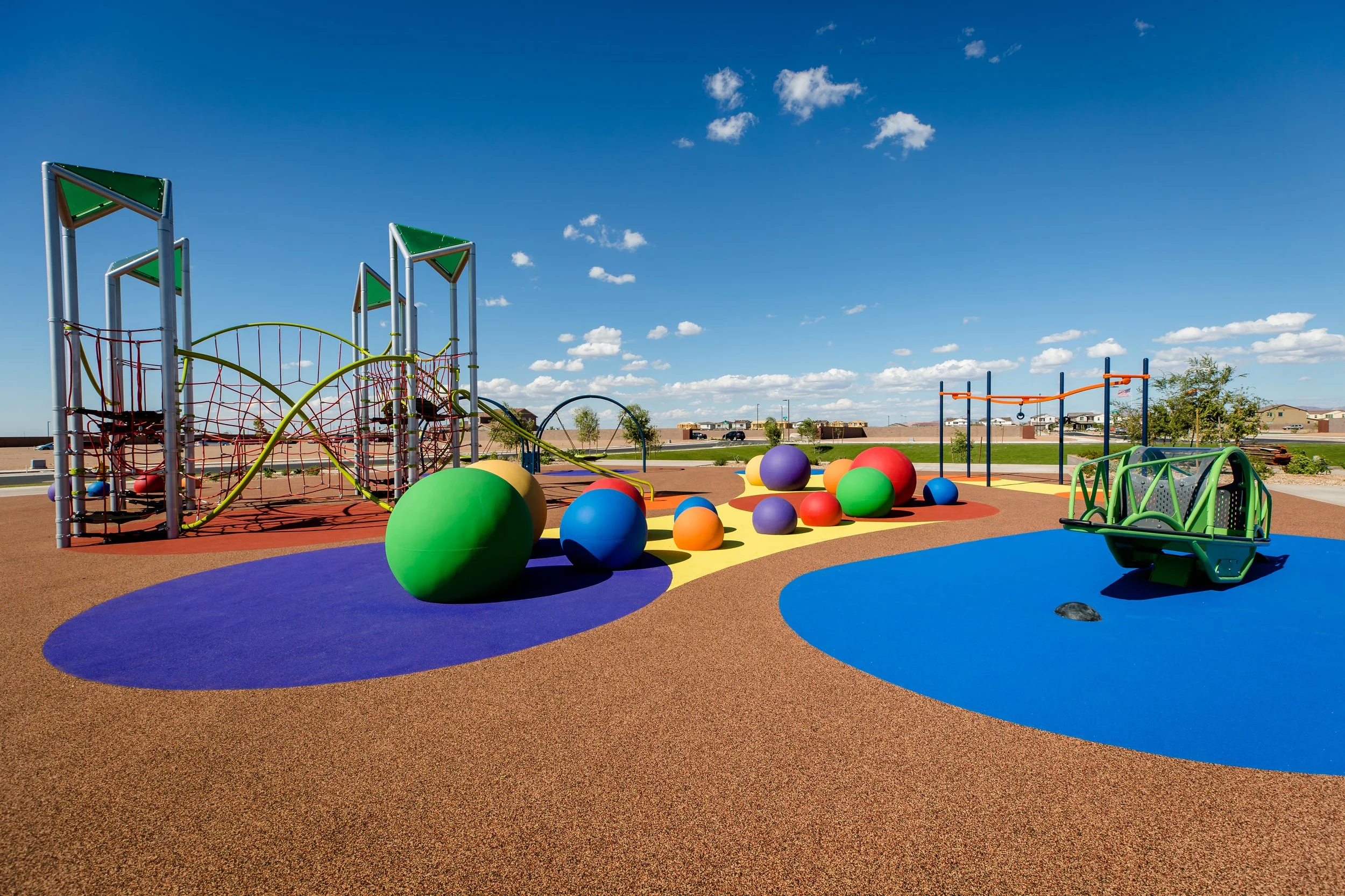 Colorful children's playground with climbing structures, large exercise balls, and swing equipment under a blue sky with clouds.