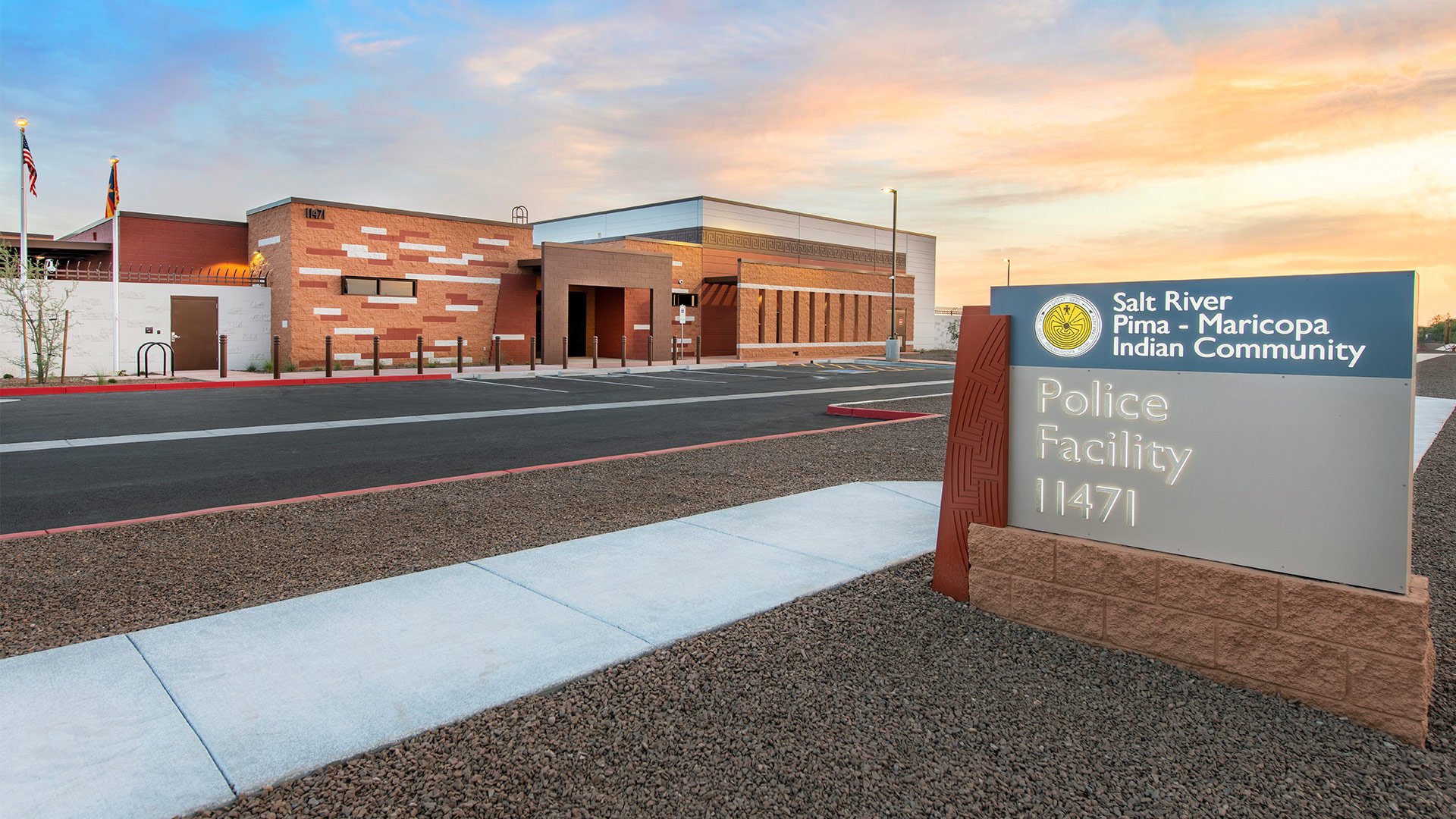 The image shows a building at sunset with a sign in the foreground that reads 'Salt River Pima-Maricopa Indian Community Police Facility 1471.' The building has a modern design with a combination of brick and white walls, and multiple flags are displ