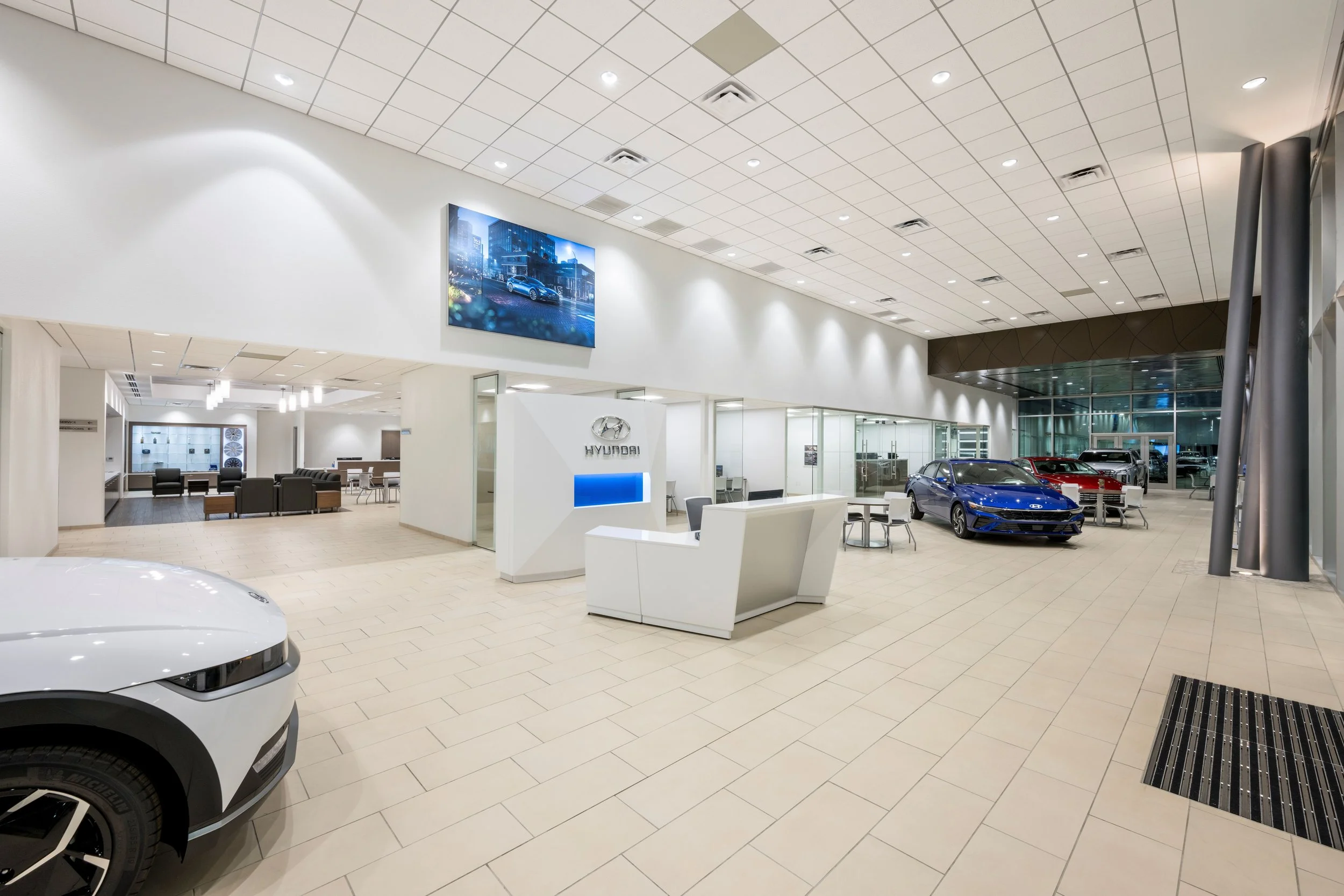 The image shows the interior of a modern Hyundai car dealership, with several cars on display, including a blue one in the center. There is a reception desk with a Hyundai logo, seating areas, and a large digital screen on the wall.