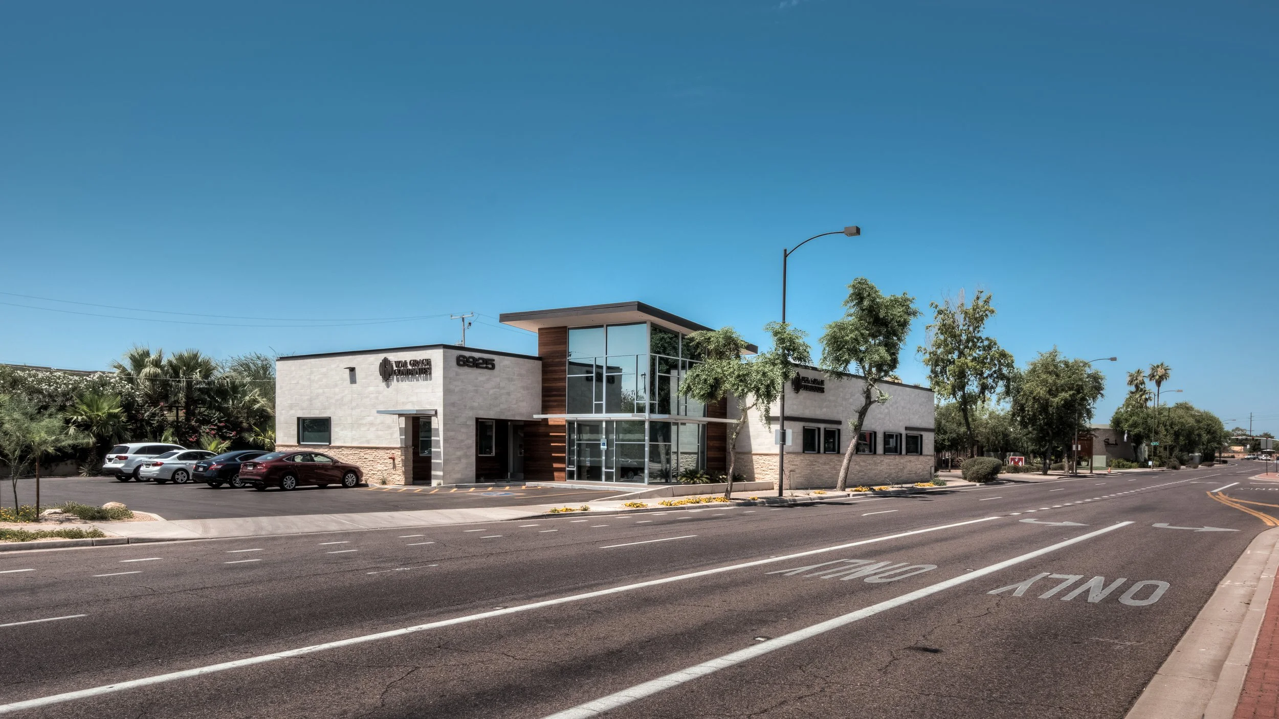 Modern commercial building with glass entrance, surrounded by trees and parked cars, along a street with road markings and streetlights, under clear blue sky.