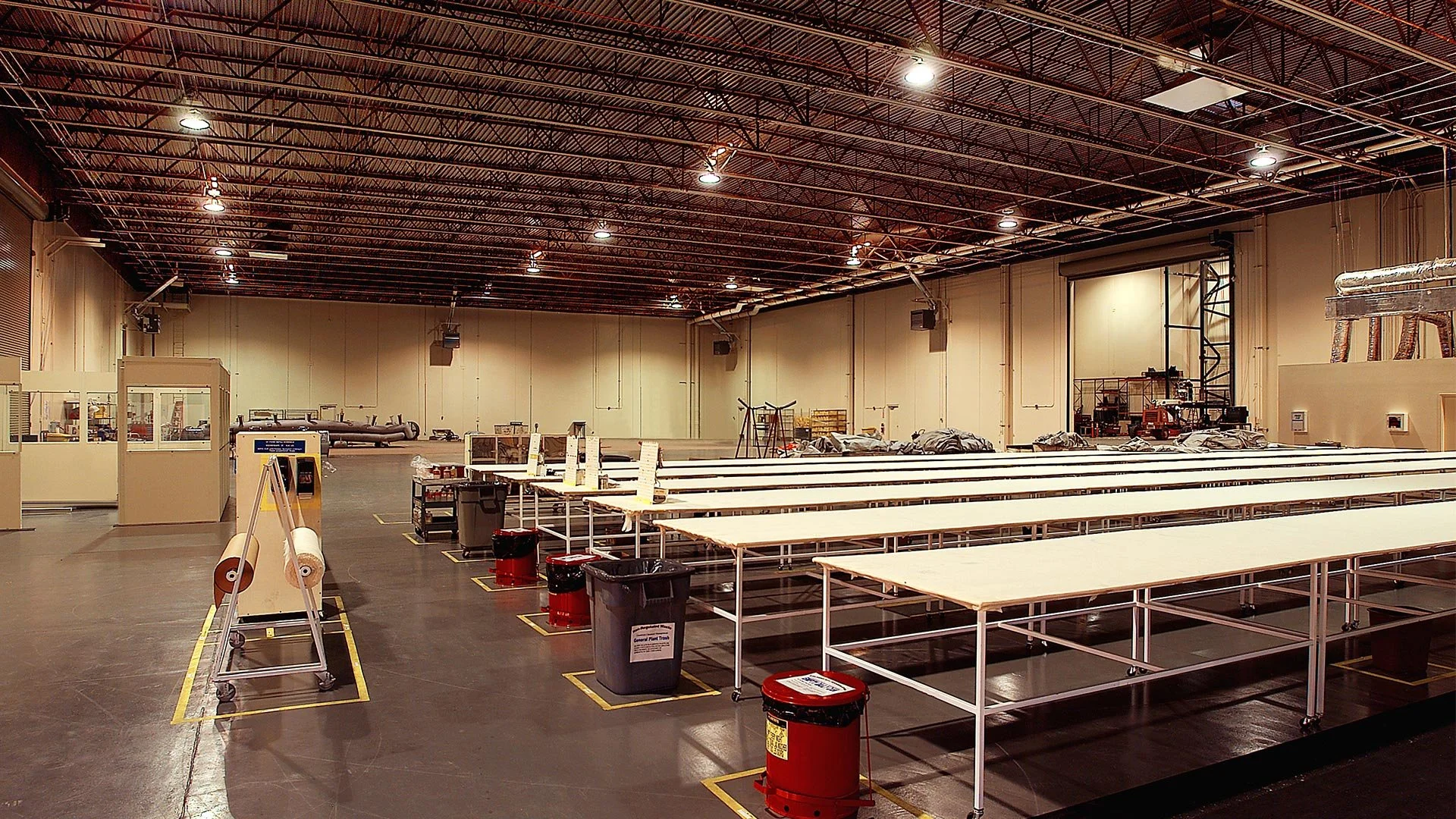 Empty industrial or commercial space with long white tables, trash bins, and equipment, possibly a cafeteria or workspace under construction or renovation.