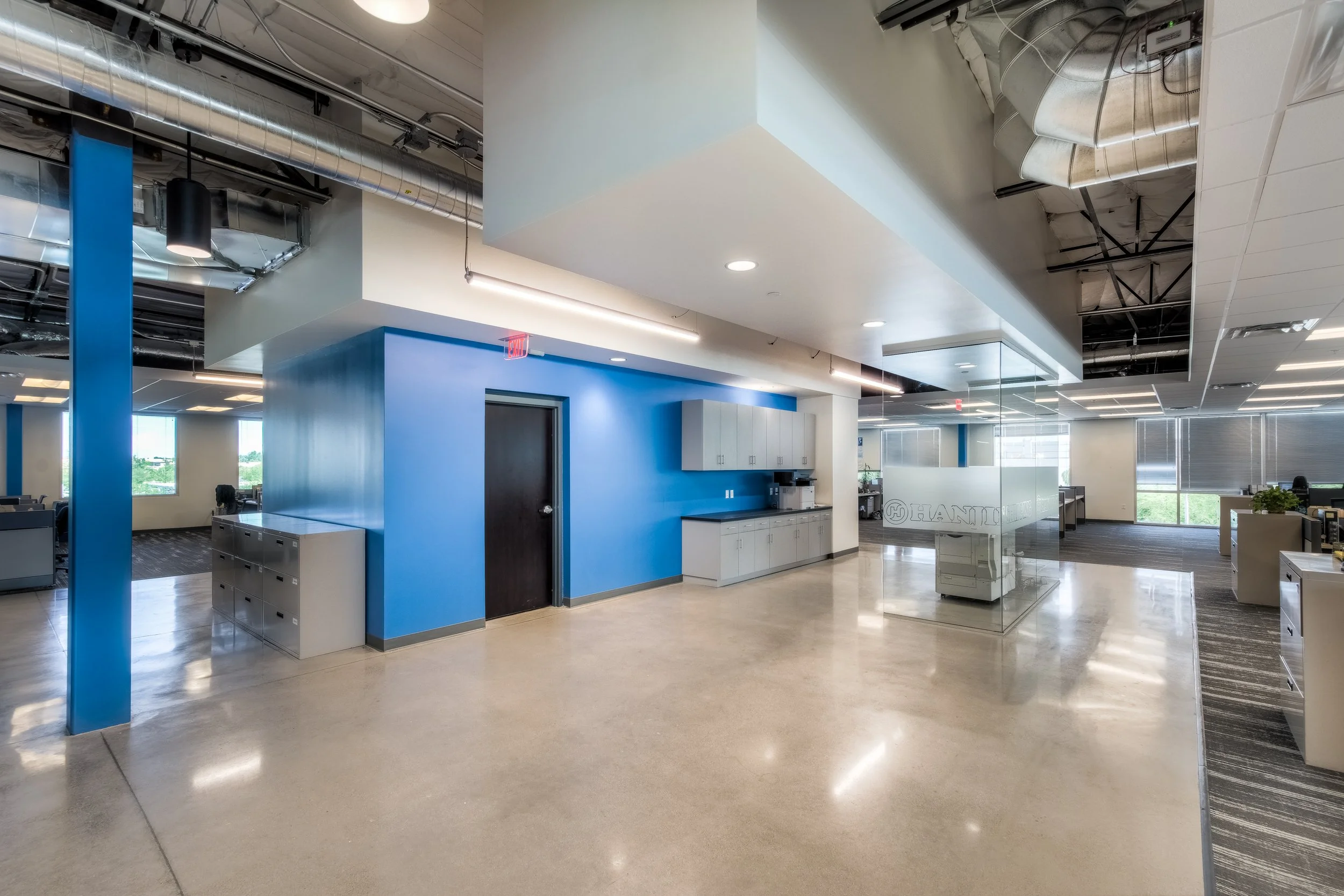 Modern office interior with blue accent wall, storage cabinets, glass partition, and open workspace with desks and windows.