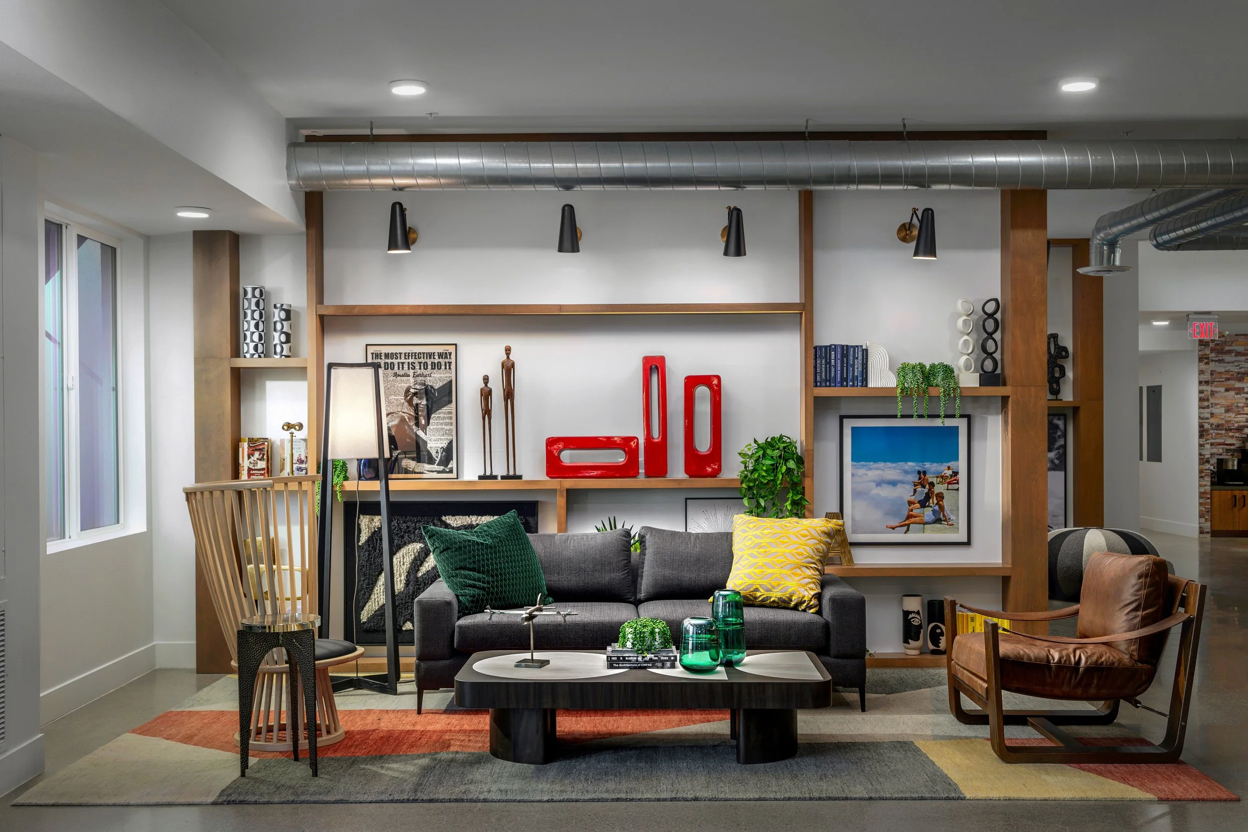 Living room with gray sofa, leather armchair, coffee table, colorful rug, and decorative wall shelves with art, books, plants, and sculptures.