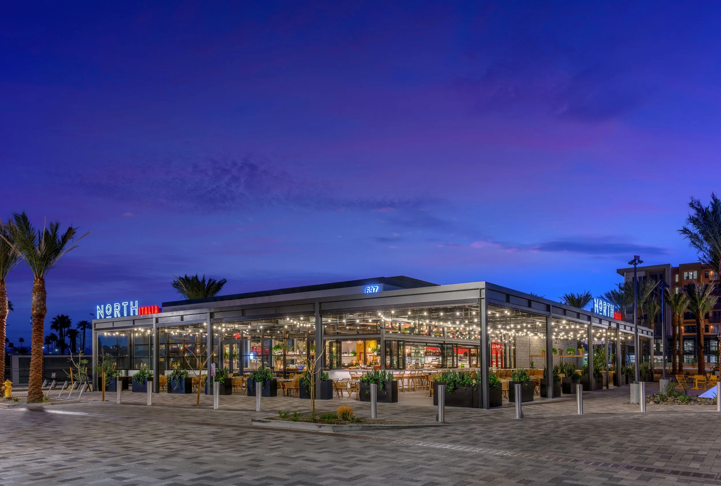 A modern outdoor dining restaurant with glass walls, string lights, and outdoor seating, surrounded by palm trees under a twilight sky.