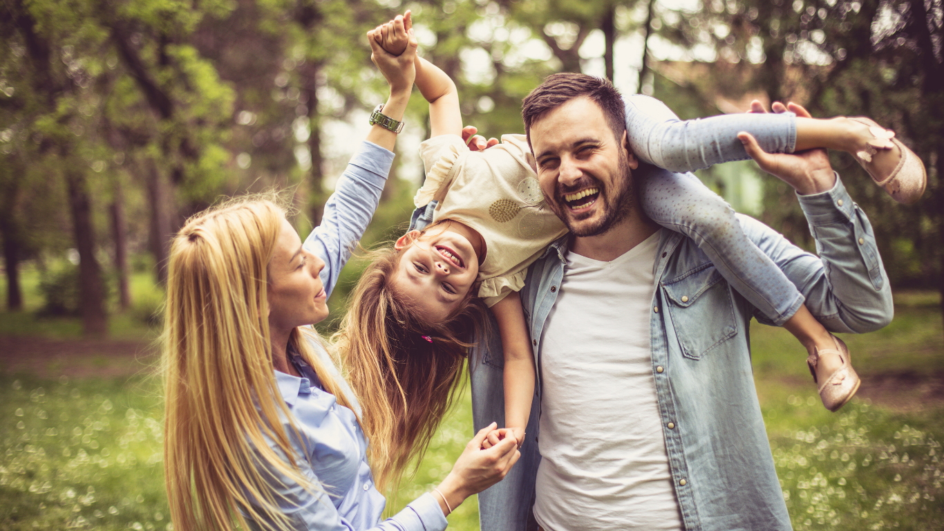 A family enjoying time together outdoors in a forest, with a man carrying a smiling girl on his shoulder and a woman holding hands with her.
