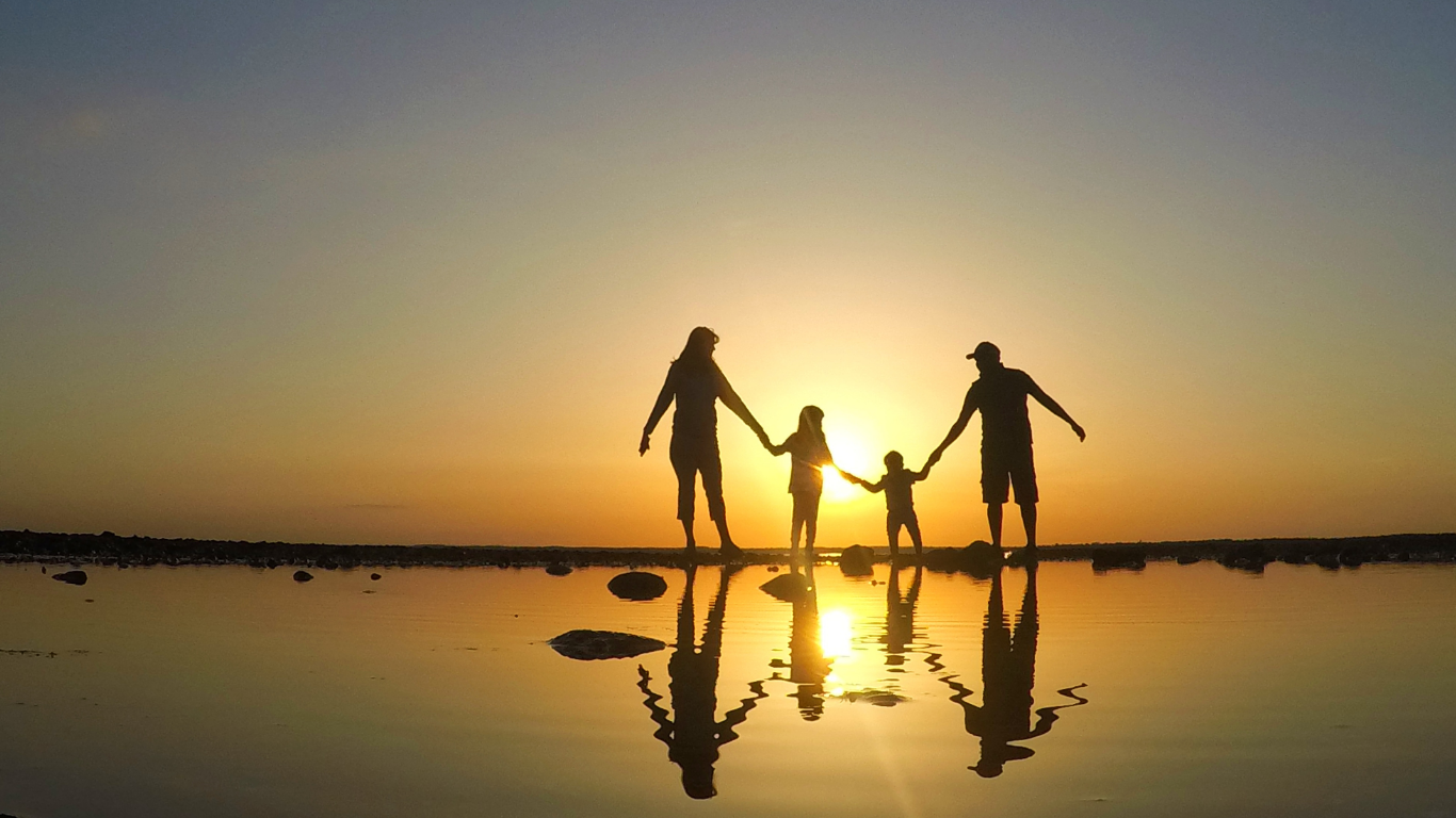 Silhouetted family of four holding hands at sunset on a beach, reflected in the water.
