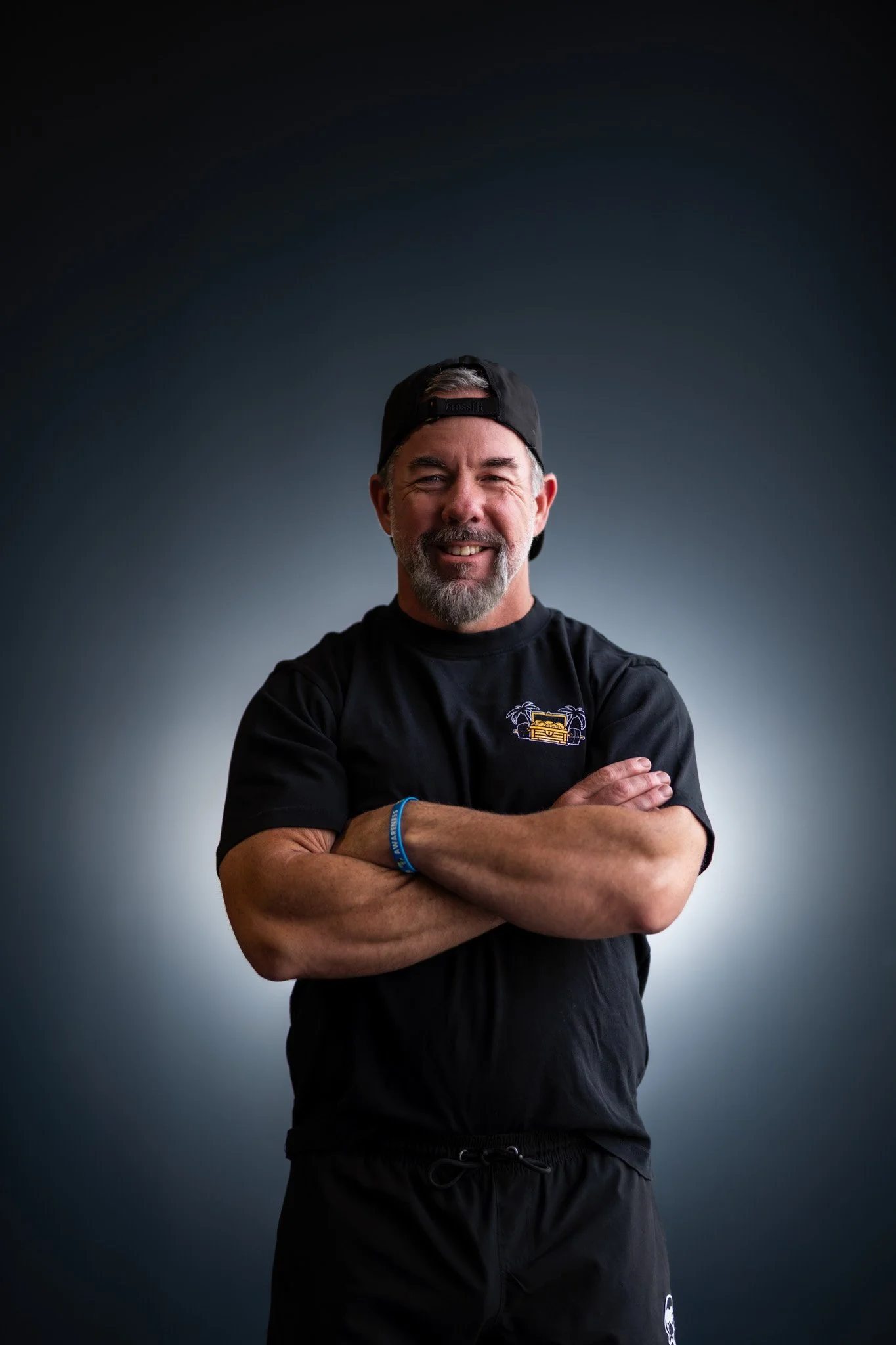 A smiling middle-aged man with a beard, wearing a black cap backwards, a black T-shirt with a small graphic on the chest, and black shorts, standing with arms crossed against a dark background.