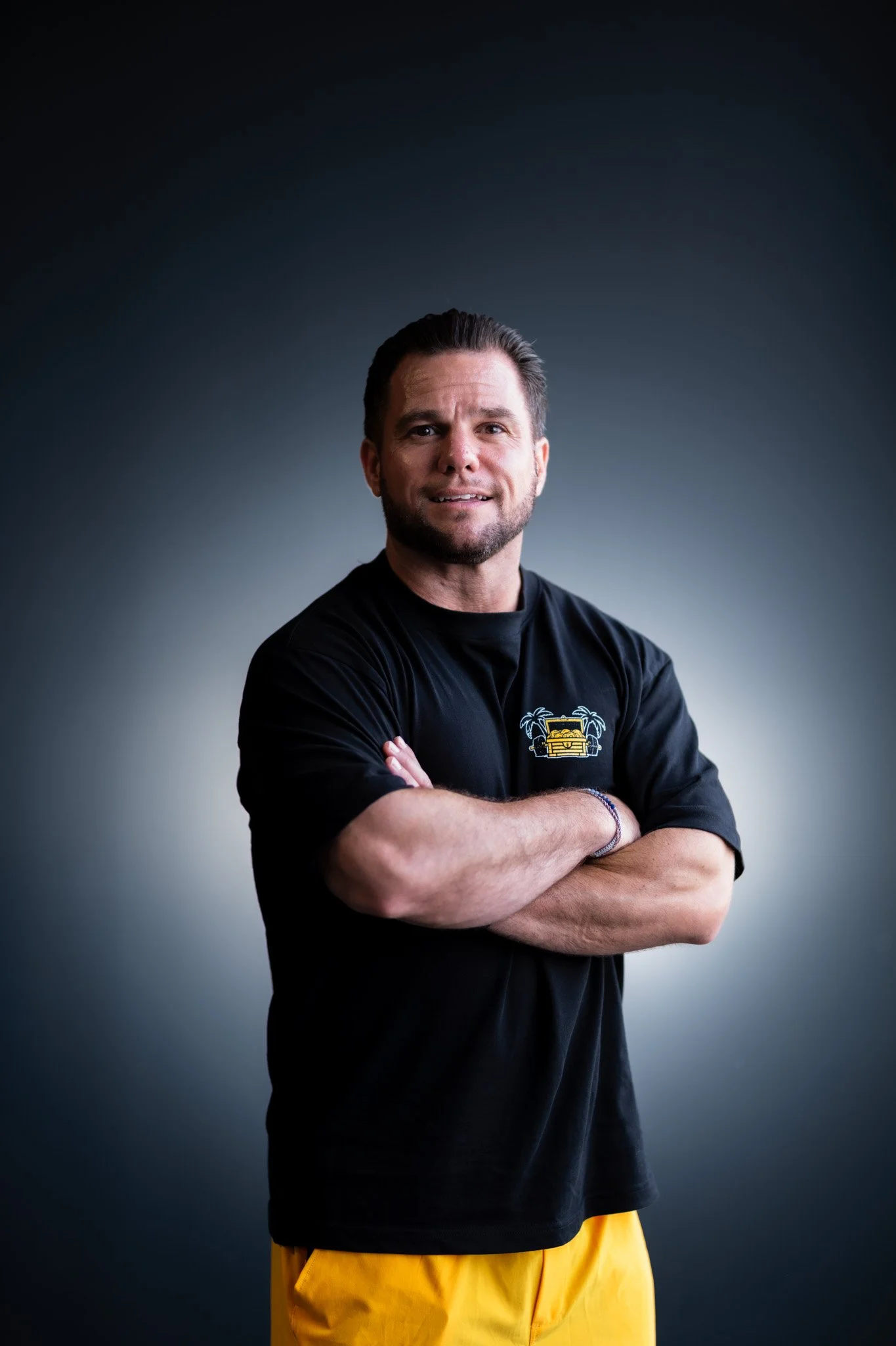 A man with dark hair and a beard, crossing his arms, wearing a black T-shirt with a small graphic on the left chest and yellow shorts, standing against a dark gradient background.