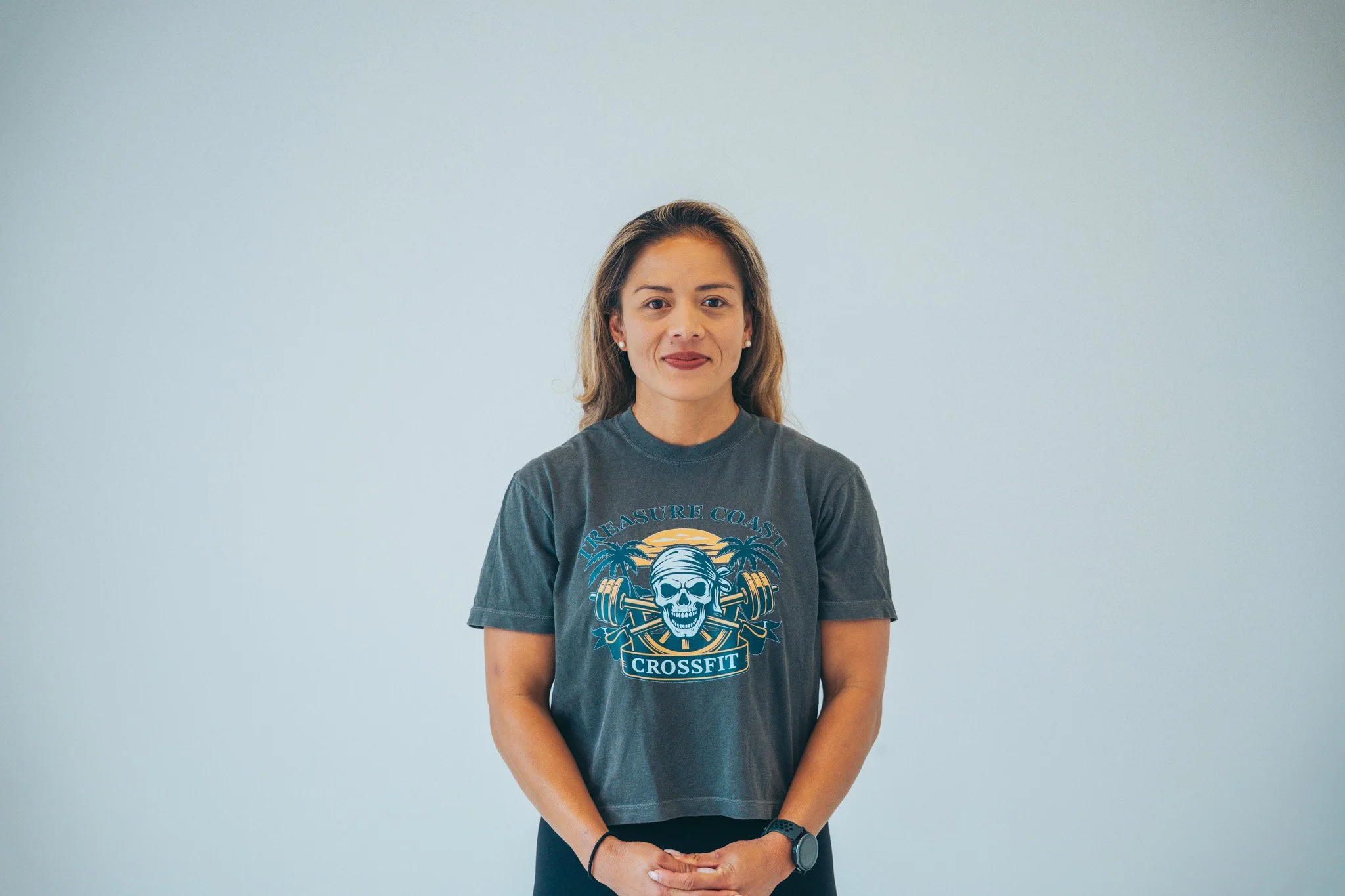 A young woman standing against a plain white background, wearing a dark grey T-shirt with a skull and crossfit design and the words "Treasure Coast" and "Crossfit" on it, with her hands clasped in front of her.
