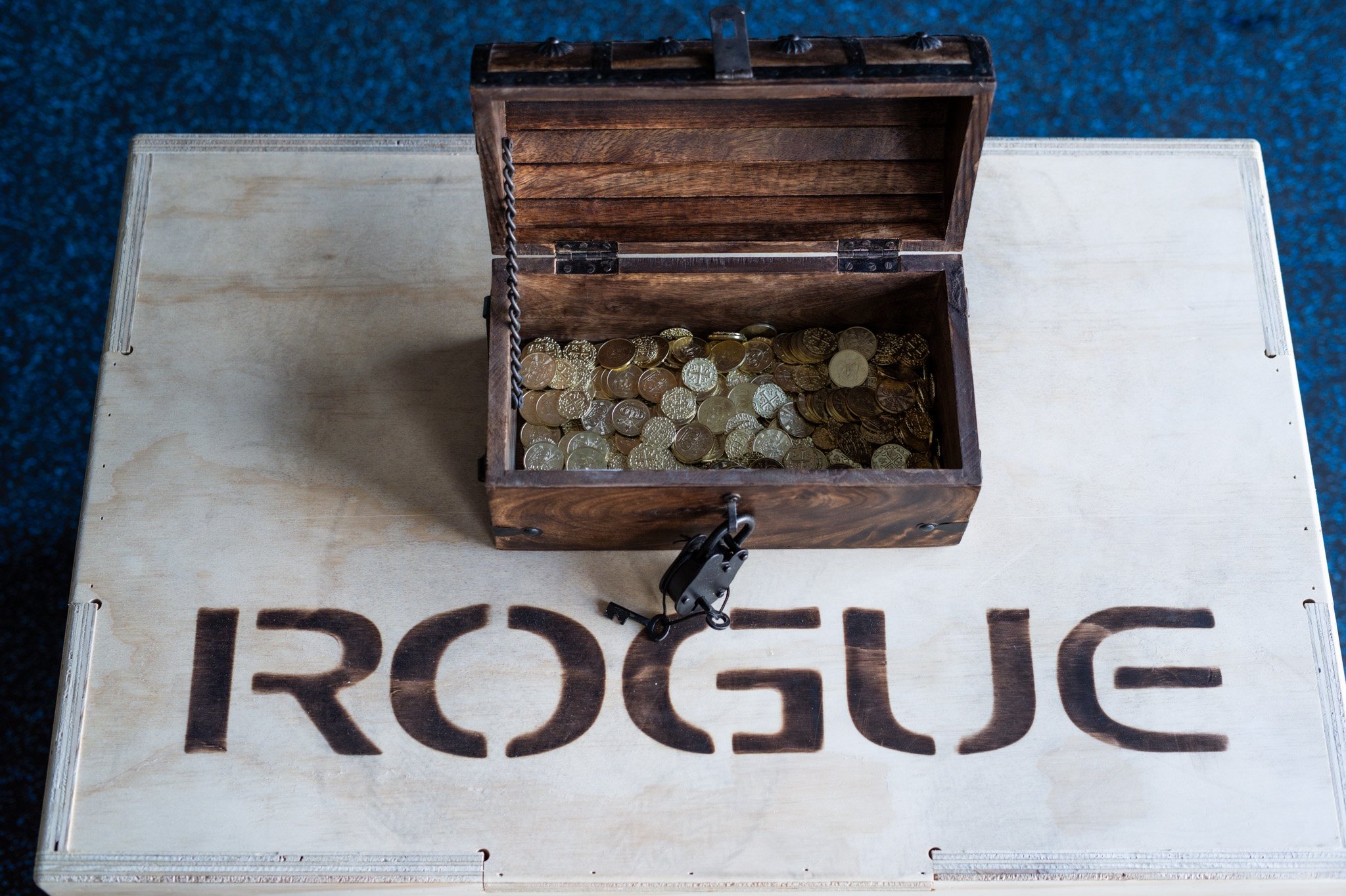 An open wooden treasure chest filled with coins sits on a wooden board with the word 'ROGUE' burned into it.