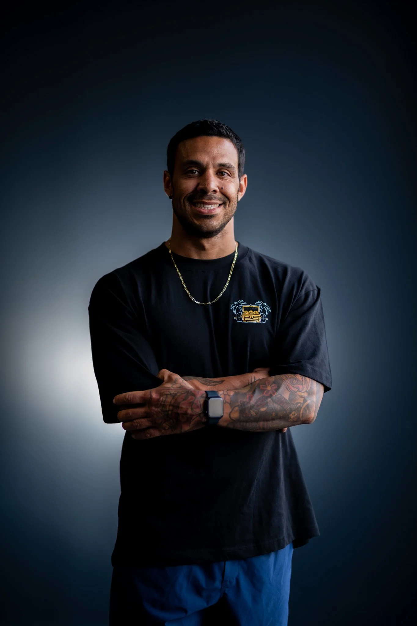 A man with tattoos on his arms, wearing a black T-shirt with a yellow graphic, a gold chain, and a smartwatch, smiling with arms crossed against a plain dark gradient background.