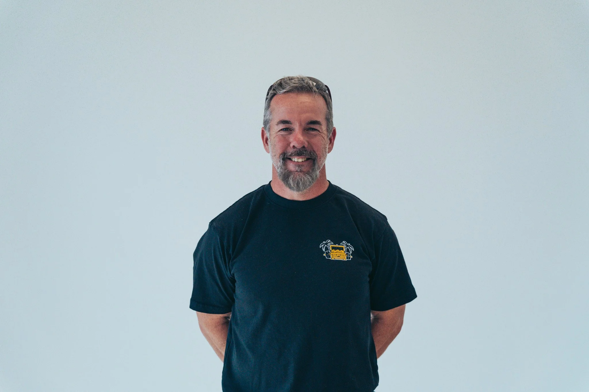 A middle-aged man with gray hair, beard, and mustache, smiling, standing against a plain light background, wearing a black T-shirt with a graphic of a yellow car, palms behind his back.