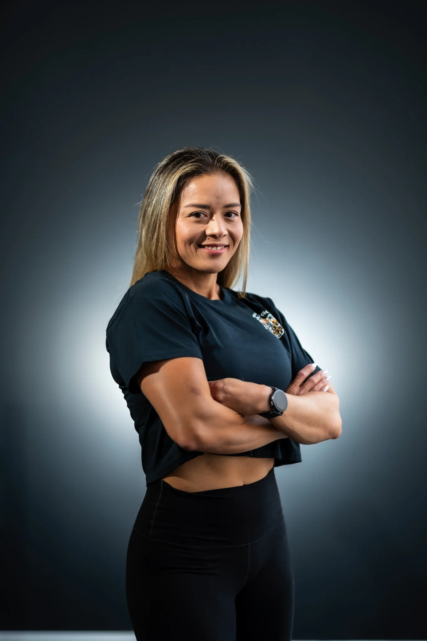 A young woman with blonde hair, smiling, is standing with her arms crossed against a dark gradient background. She is wearing a black t-shirt with a logo on it and black leggings, with a smartwatch on her left wrist.