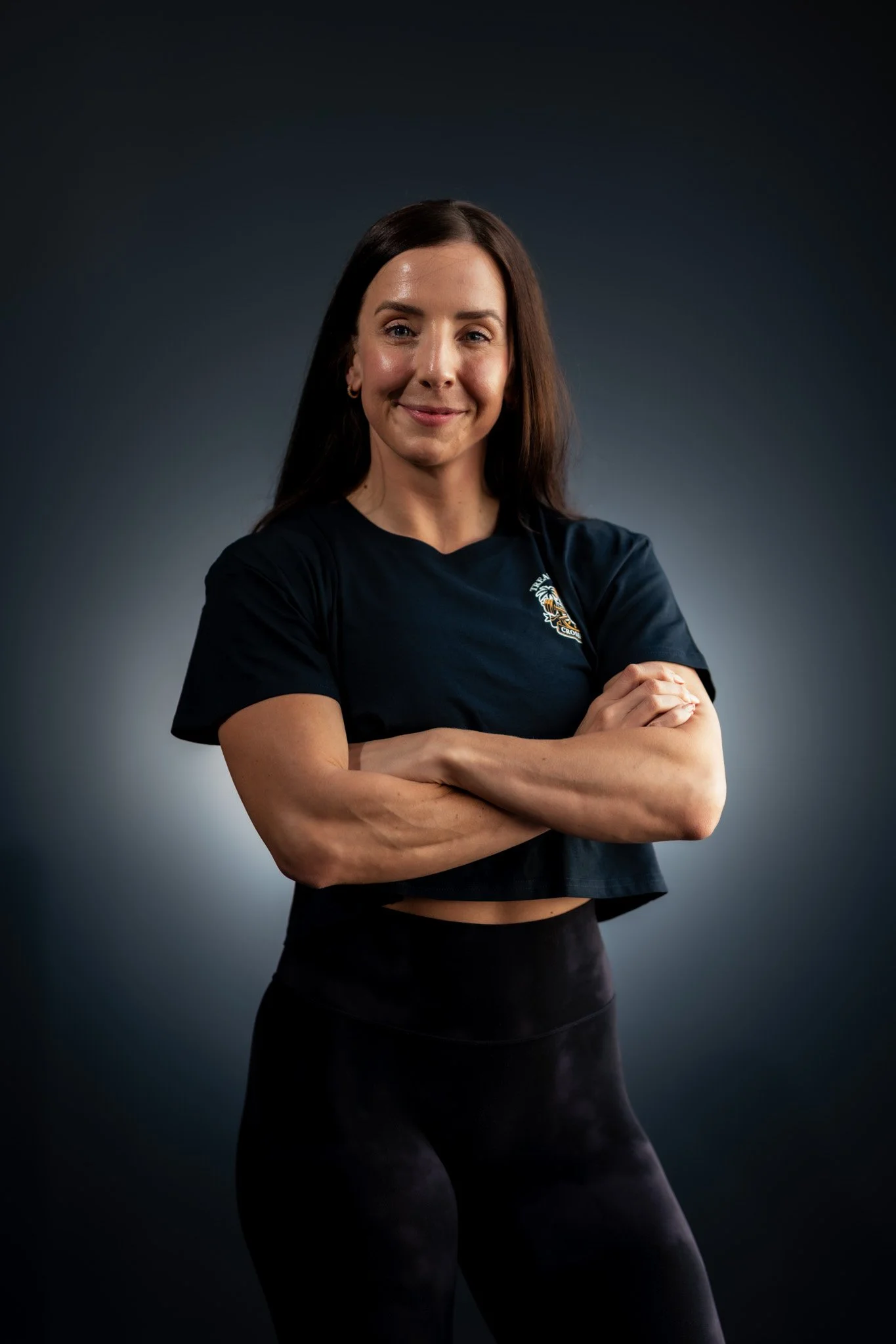 A woman with long brown hair, smiling, wearing a black crop top T-shirt and black leggings, standing with arms crossed against a dark background.