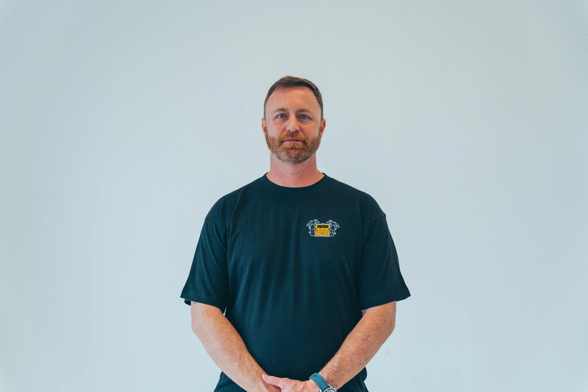 A man with a beard and short hair standing against a plain light-colored background, wearing a navy blue T-shirt with a small yellow graphic of a couch with palm trees on the front.