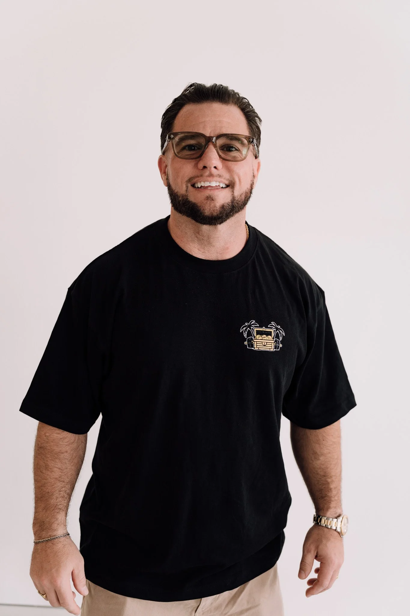 A smiling man with dark hair and beard, wearing sunglasses, a black T-shirt with a palm tree and vintage car graphic, and beige pants, standing against a plain white background.