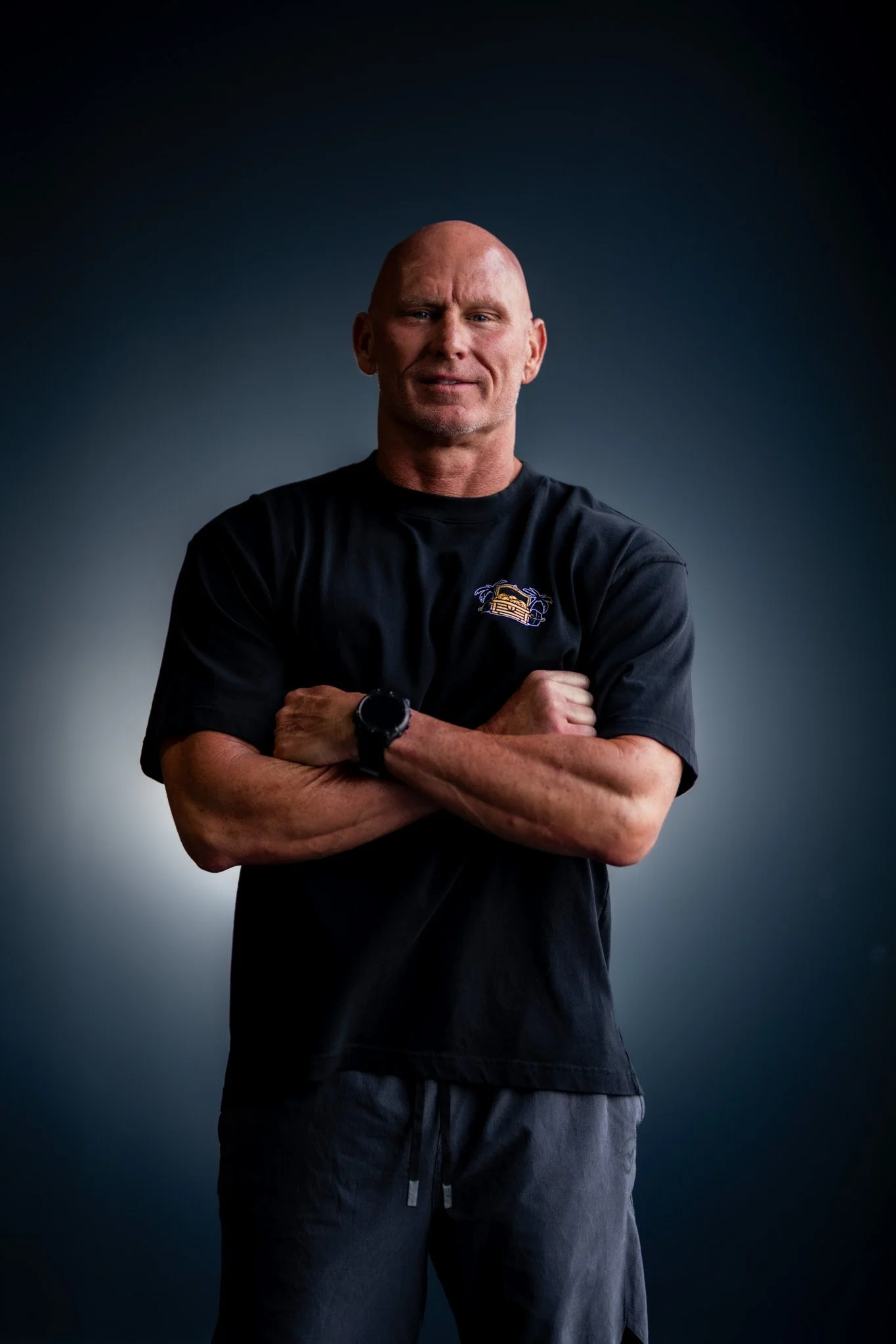 A middle-aged man with a shaved head and a slight smile, crossing his arms, wearing a black T-shirt with a small logo and a wristwatch, standing against a dark gradient background.