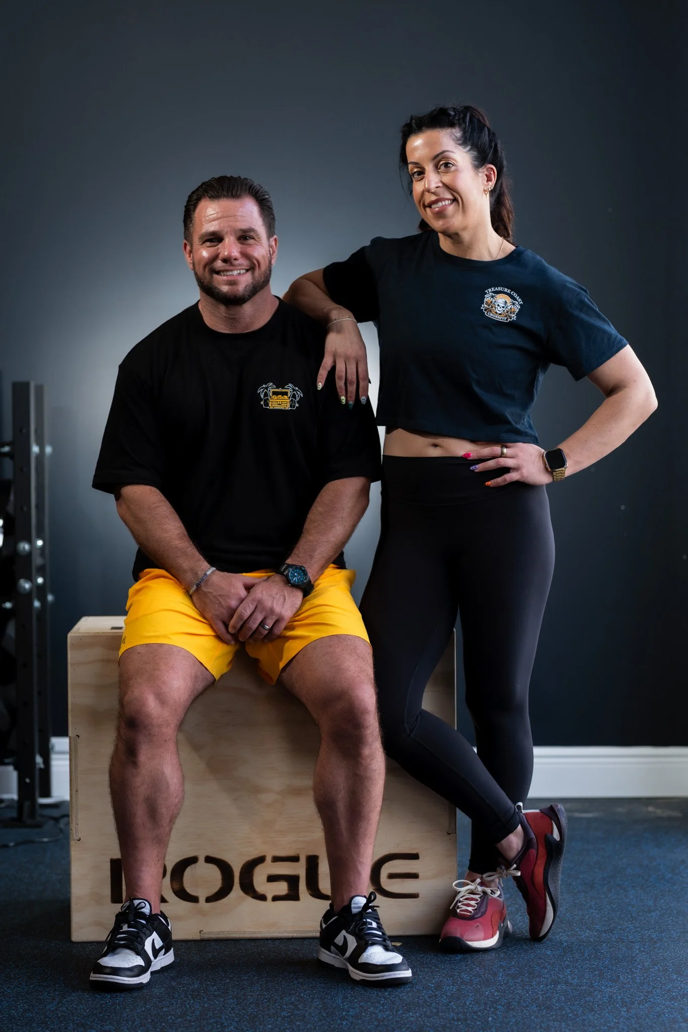 A man and a woman pose together in a fitness studio, with the man seated on a wooden plyometric box and the woman standing beside him with her hand on his shoulder. Both are dressed in casual workout clothes, smiling at the camera.