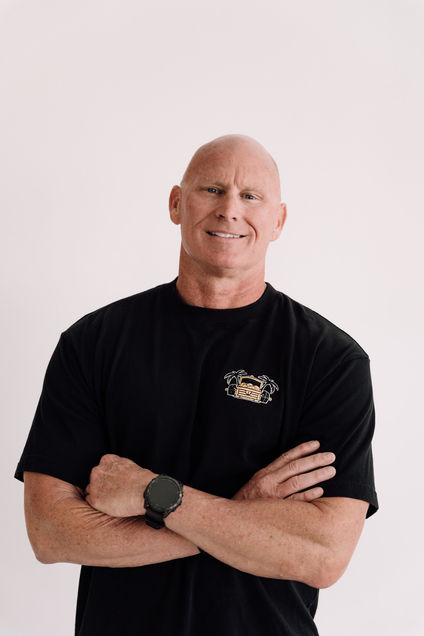 A smiling bald man with light skin, wearing a black t-shirt with a small graphic of a classic car and palm trees on the chest, stands with arms crossed against a plain white background.