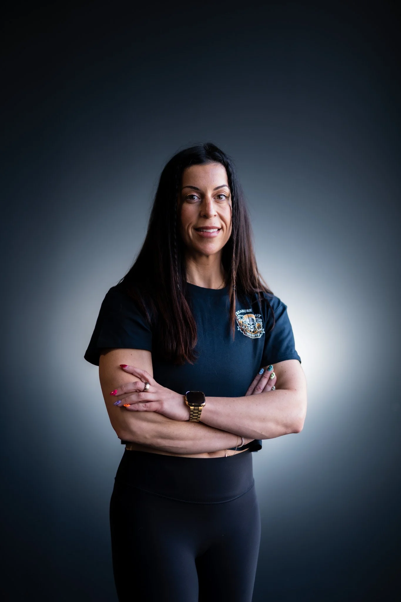 A woman with dark hair, wearing a black t-shirt with a logo, standing against a dark gradient background, arms crossed.