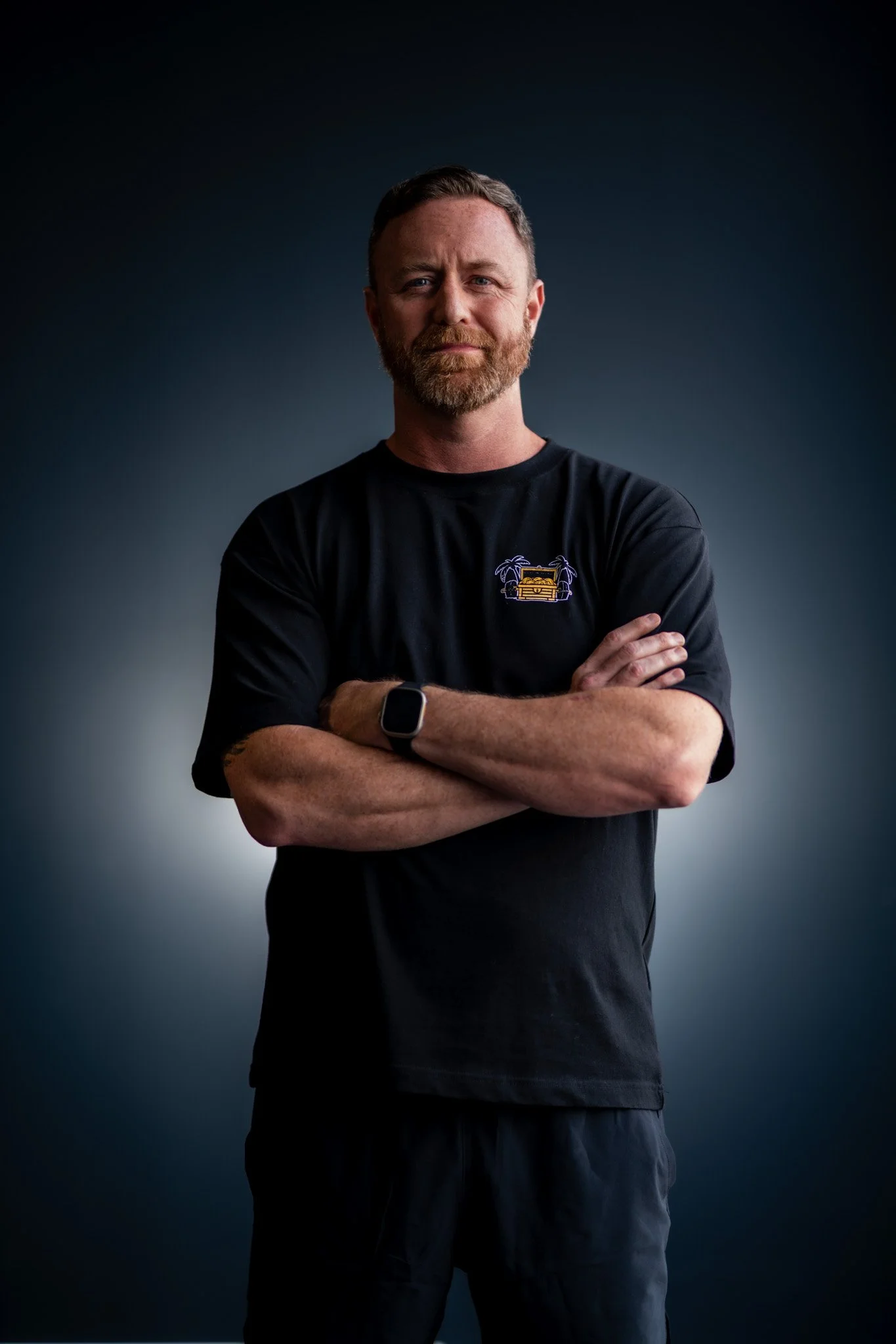 A man with a beard and mustache standing with arms crossed, wearing a black t-shirt with a small embroidered design, against a dark gradient background.