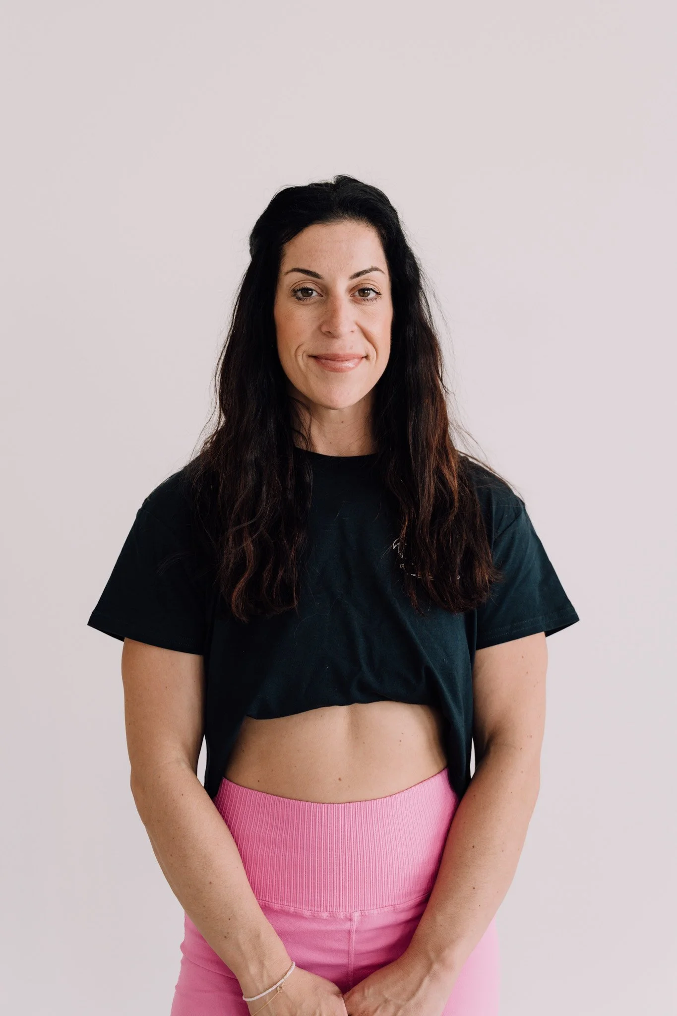A woman with long dark wavy hair, wearing a black crop top and pink high-waisted pants, standing against a plain light-colored background.