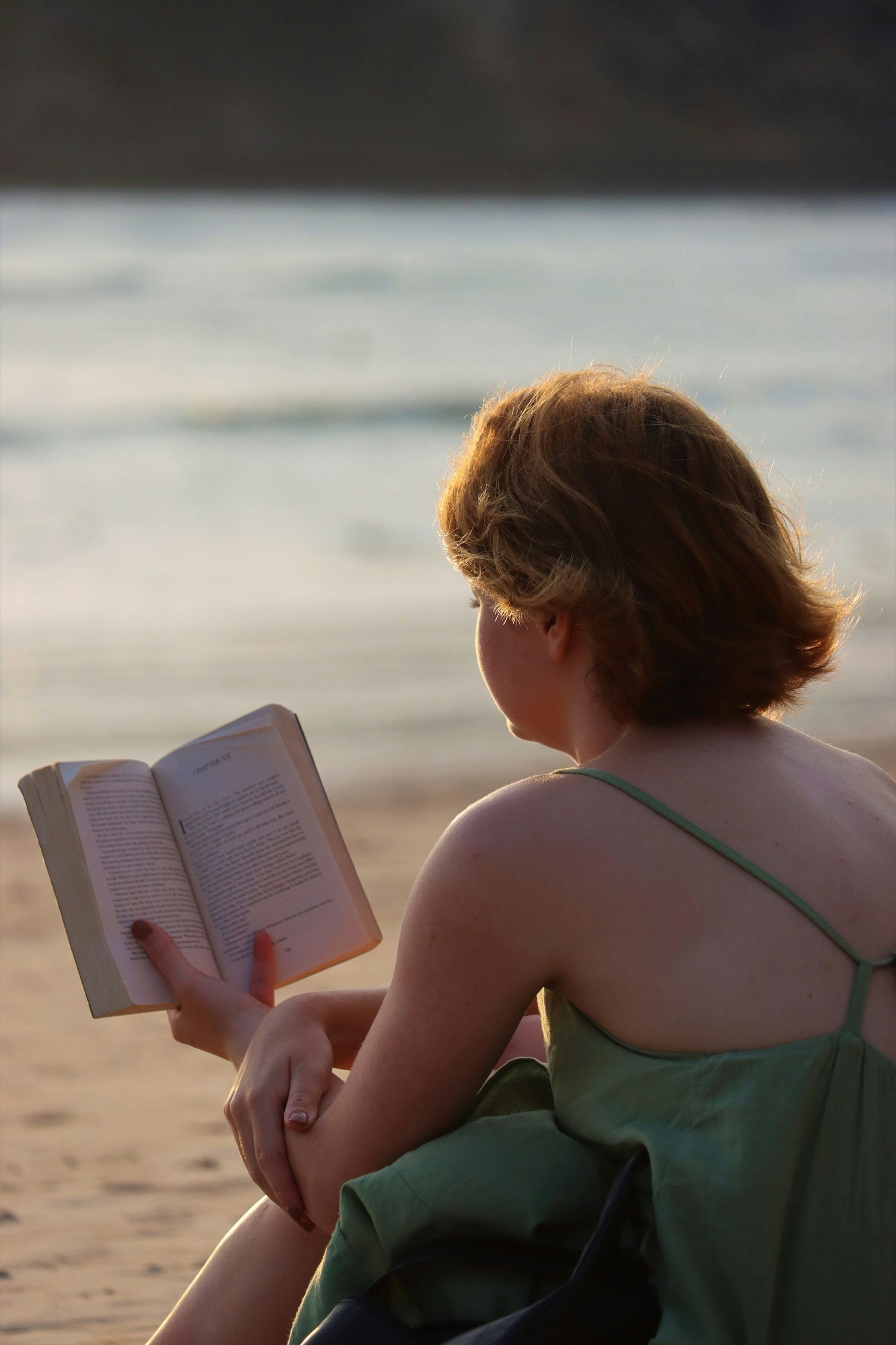 Une personne assise sur la plage, lisant un livre au coucher du soleil, avec la mer en arrière-plan.