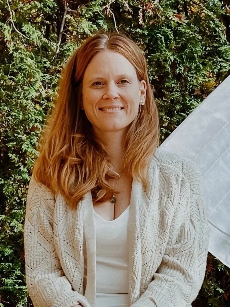 Une femme aux cheveux roux, souriante, portant un gilet beige et un t-shirt blanc, posant devant un mur de feuillage vert.