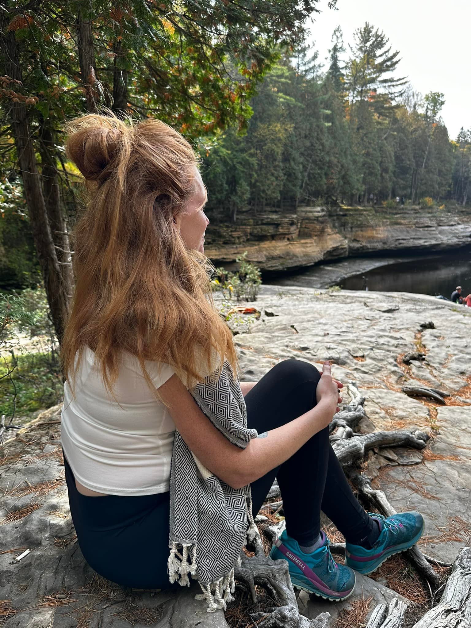 Femme aux cheveux roux, assise sur un rocher au bord d'une rivière, entourée d'arbres, portant un t-shirt blanc, un foulard, des vêtements de sport, des chaussures de randonnée, en pleine nature.