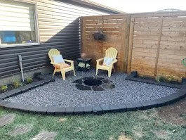 Backyard patio with two yellow Adirondack chairs and a fire pit, surrounded by gray gravel and wooden fencing.