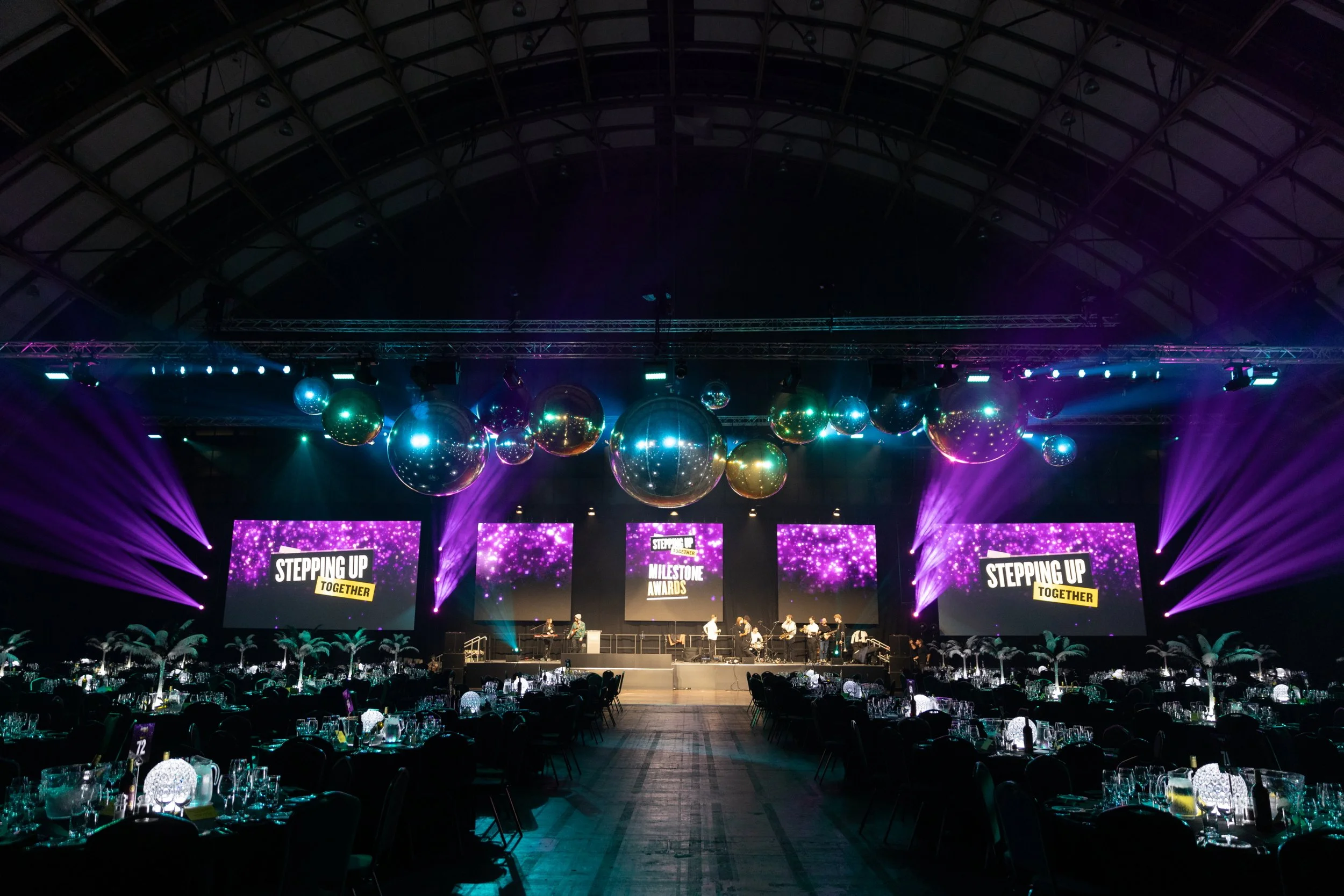 Indoor event venue with a stage, large screens displaying 'Stepping Up Together', tables with place settings and centerpieces, and decorative hanging Silver and Gold Inflatable Mirror Balls with purple and blue lighting.