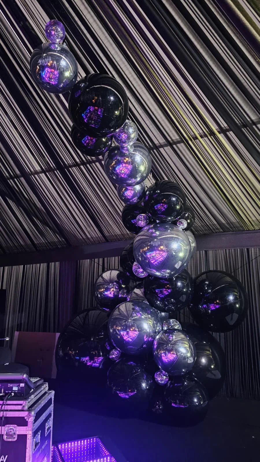 Arranged black, silver, and clear balloons with purple lighting reflections, set against a striped ceiling and wall.